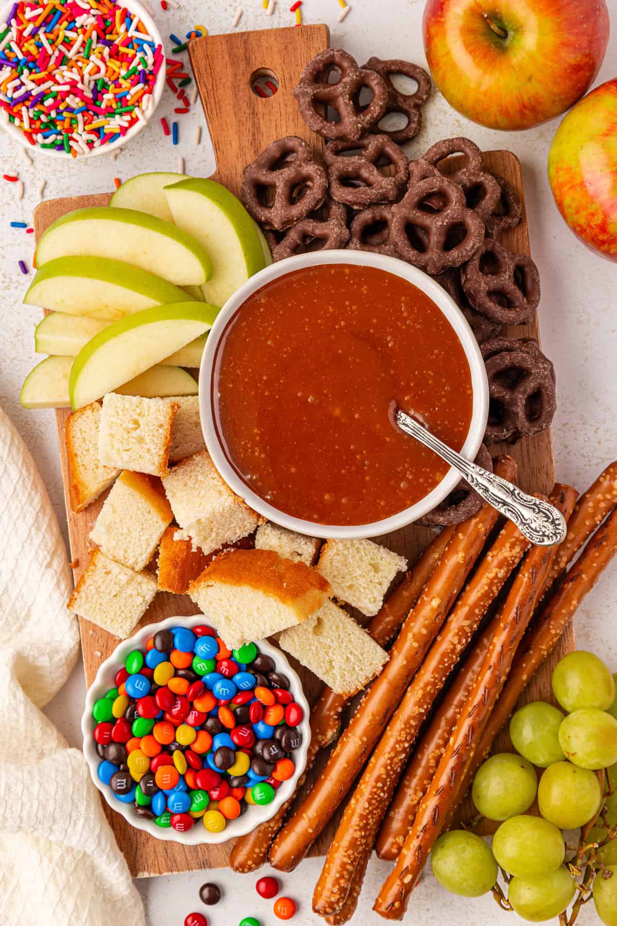 A wooden board with sliced apples, grapes, chocolate-covered pretzels, pretzel rods, cake cubes, colorful candies, and a bowl of caramel dip. Nearby are apples and a bowl of rainbow sprinkles.