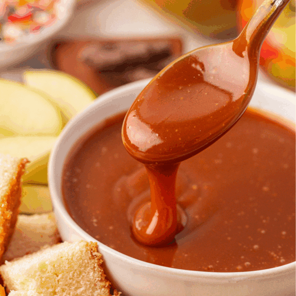 A spoon drizzles thick caramel sauce into a white bowl surrounded by slices of apple and pieces of cake.