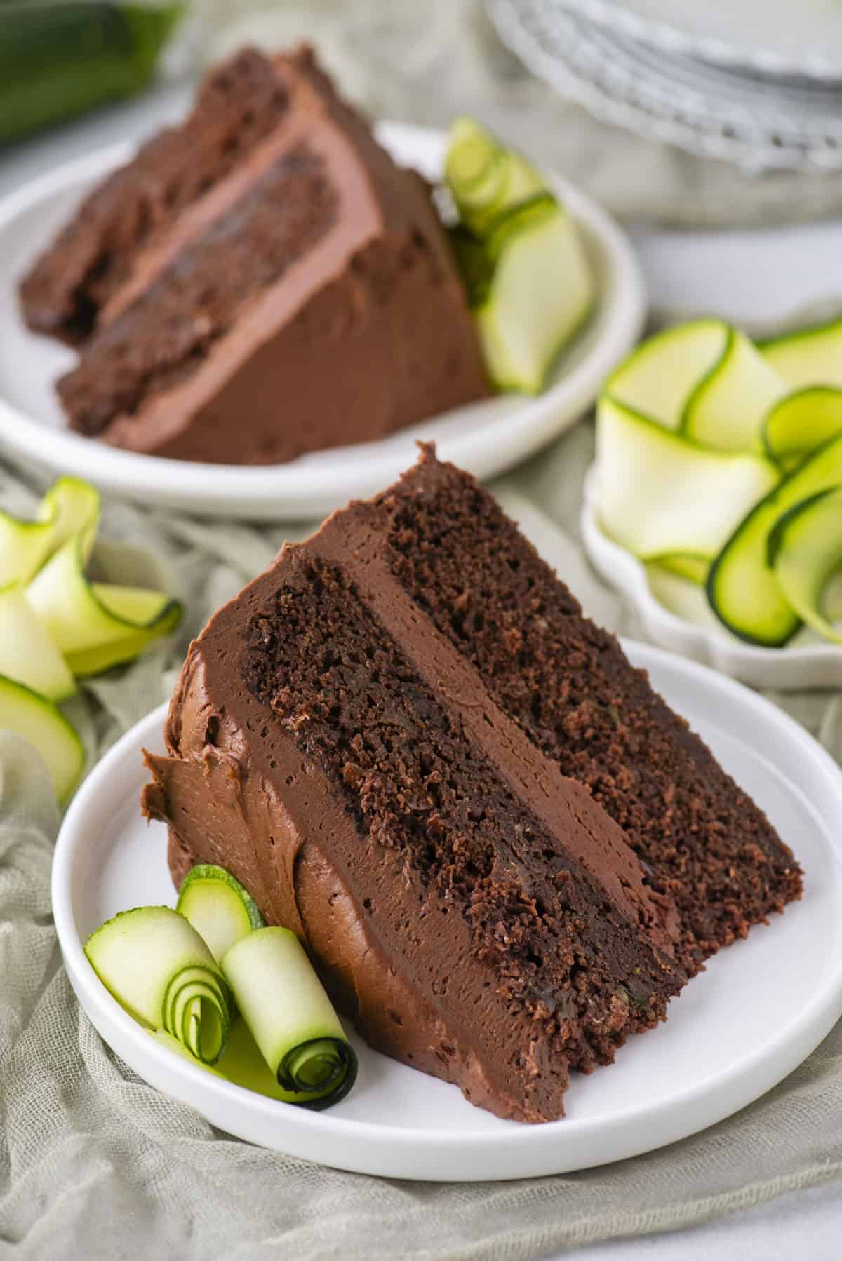 Two slices of rich chocolate cake with chocolate frosting are served on white plates, each garnished with thinly sliced, rolled zucchini ribbons. A stack of plates is blurred in the background.
