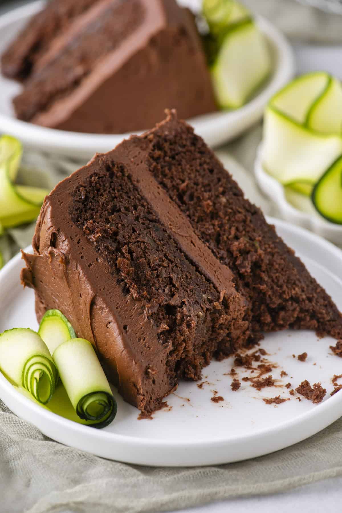 A slice of rich chocolate cake with chocolate frosting on a white plate, garnished with rolled slices of zucchini, with another plate of cake in the background.