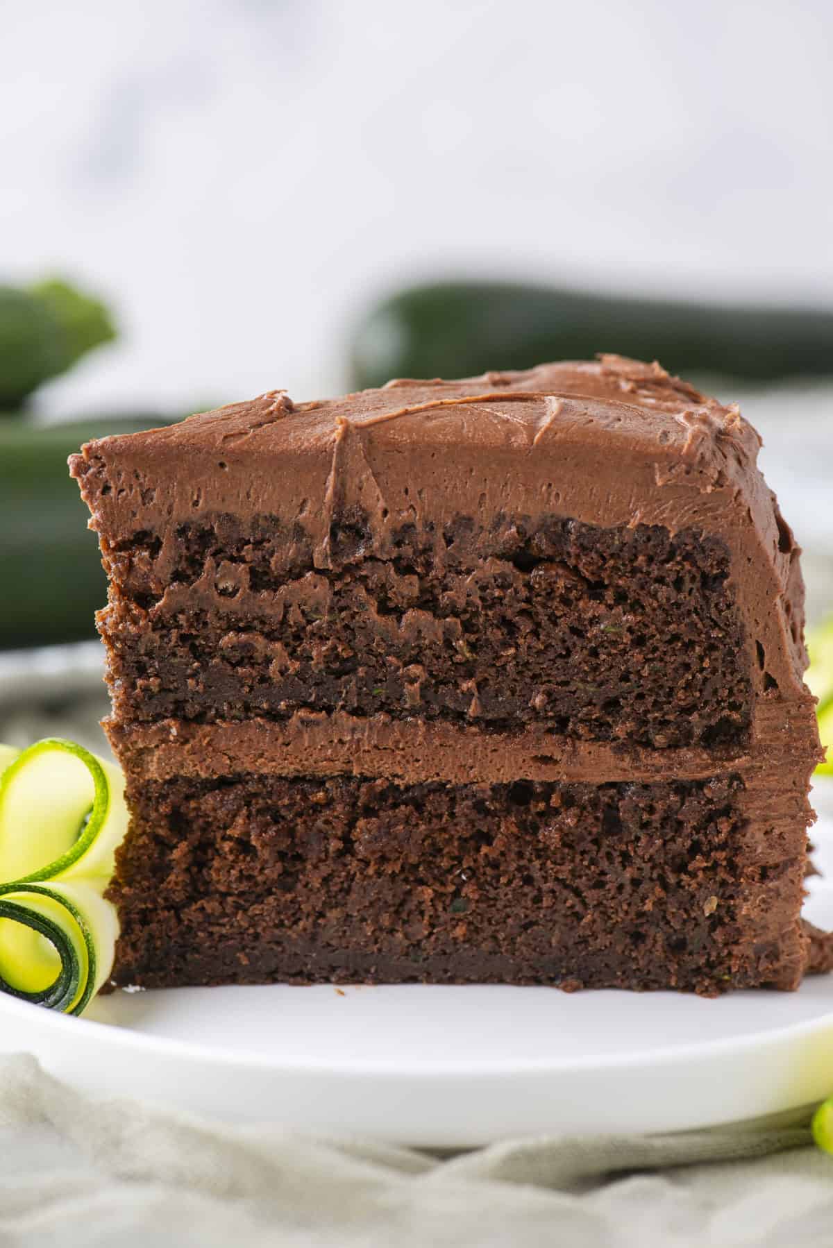A close-up of a slice of chocolate layer cake with chocolate frosting on a white plate, garnished with a curl of yellow and green zucchini. The background is softly blurred.