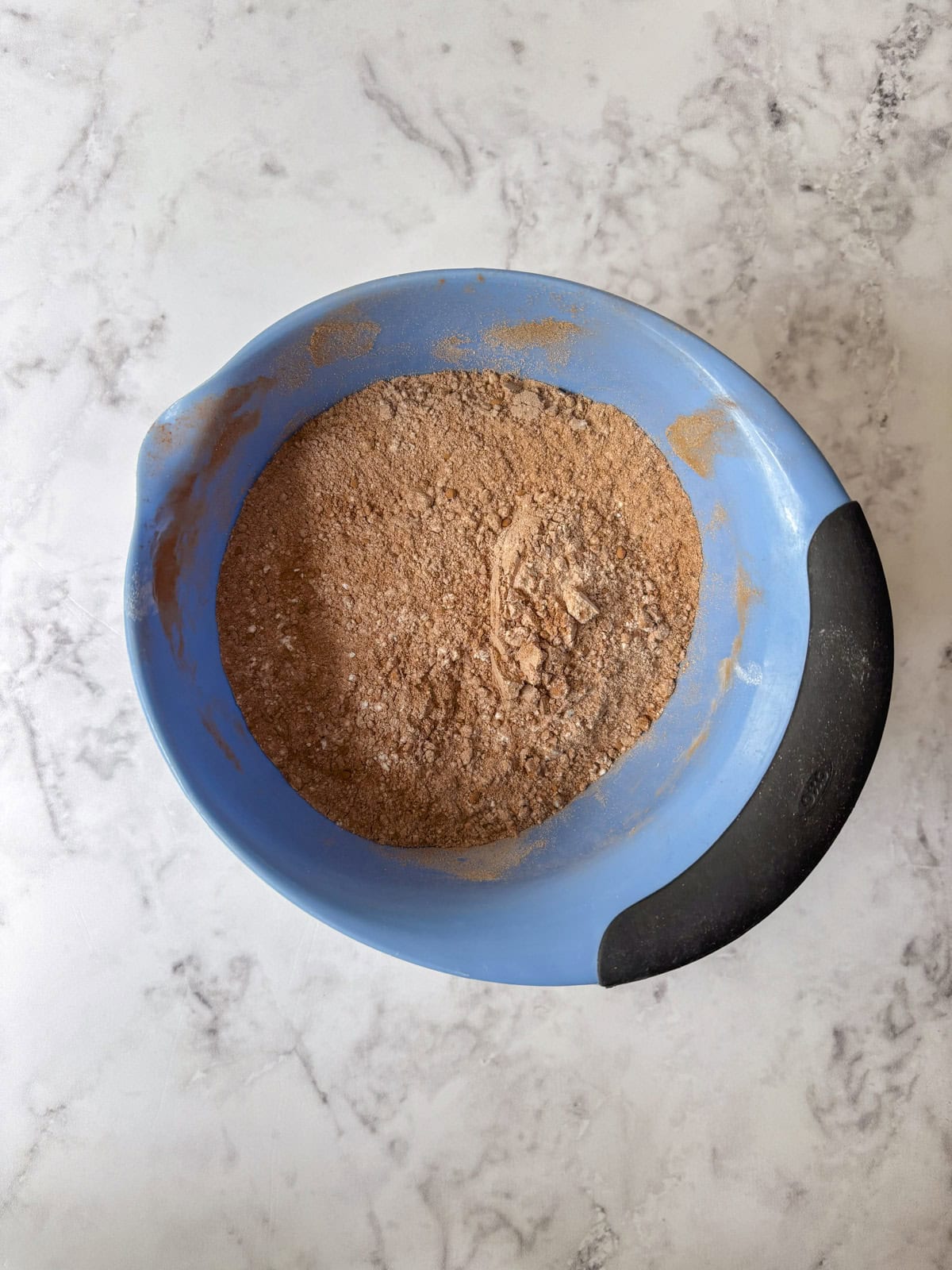 A blue mixing bowl with a black handle contains a blended mixture of dry cocoa powder and flour on a white marble surface.