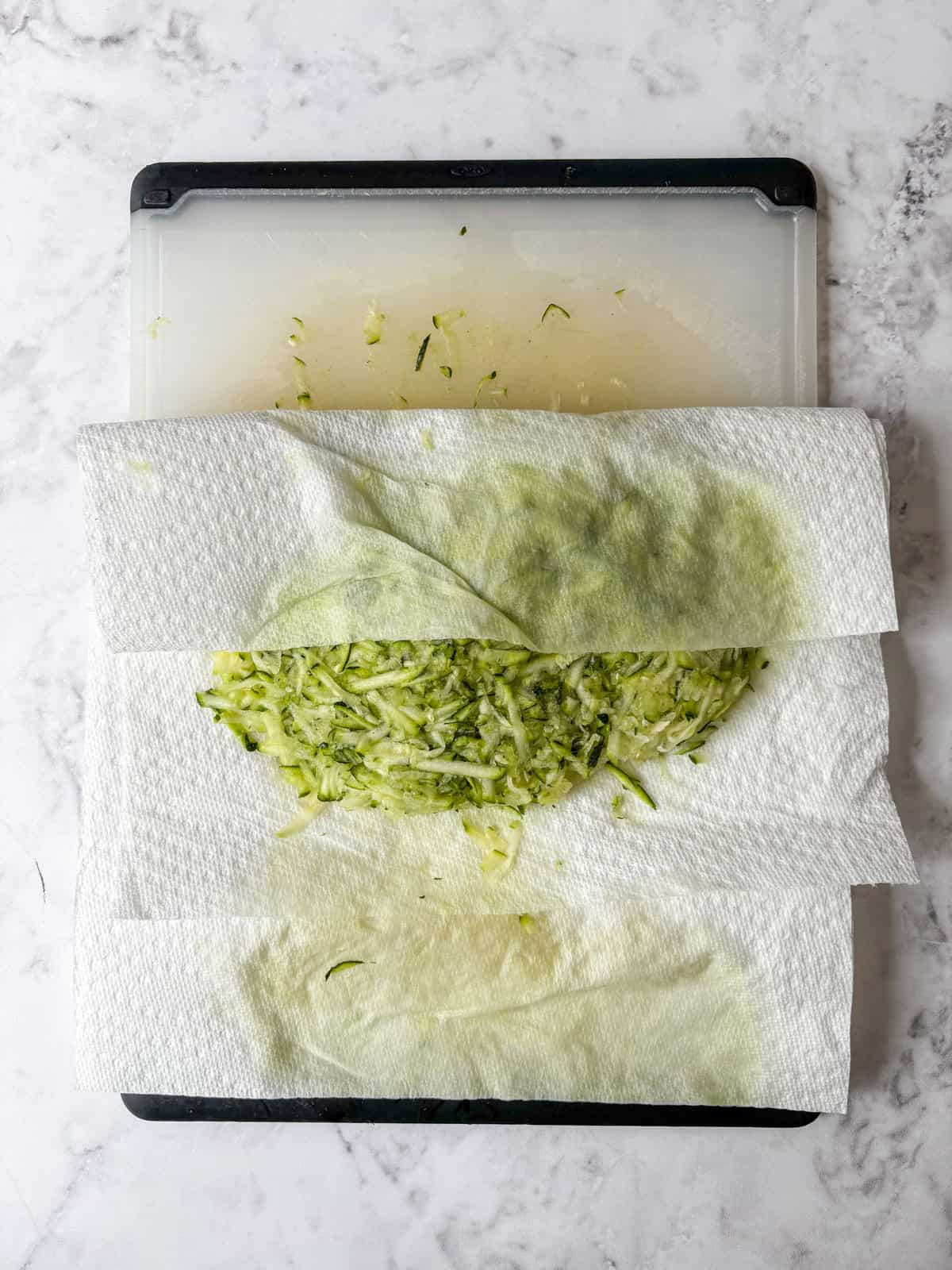 Shredded zucchini on a cutting board is partially wrapped in paper towels, with visible moisture absorbed into the towels, all sitting on a white marble surface.