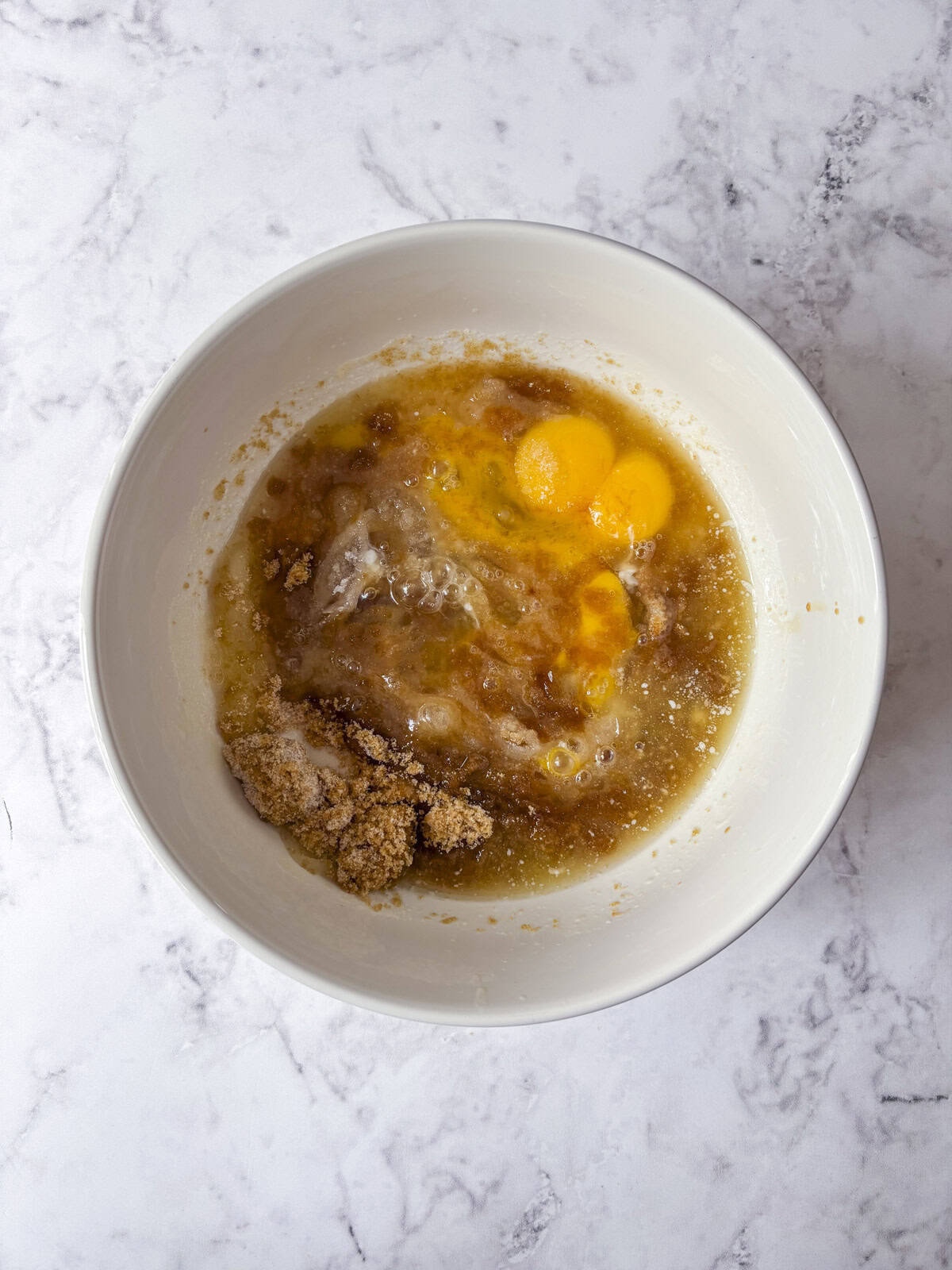 A white bowl on a marble surface containing eggs, brown sugar, and wet ingredients about to be mixed together.