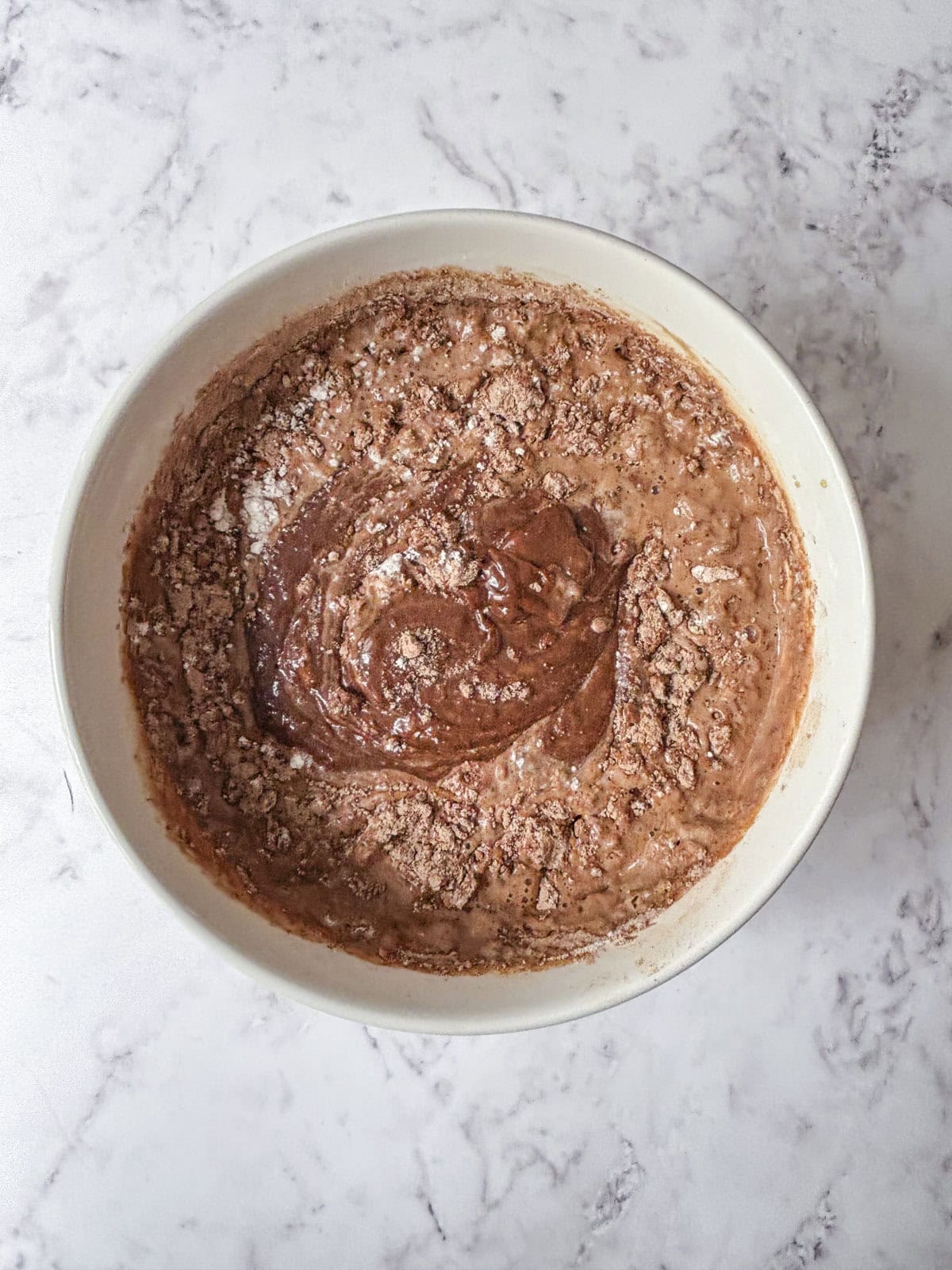 A white bowl filled with partially mixed chocolate cake batter sits on a light marble surface. Dry cocoa powder and flour are still visible on top of the wet ingredients.