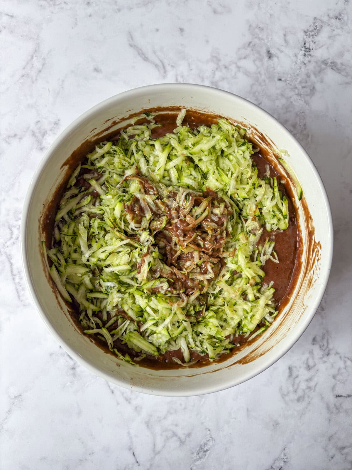 Overhead view of a mixing bowl with shredded zucchini being mixed into chocolate cake batter on a light marble countertop.