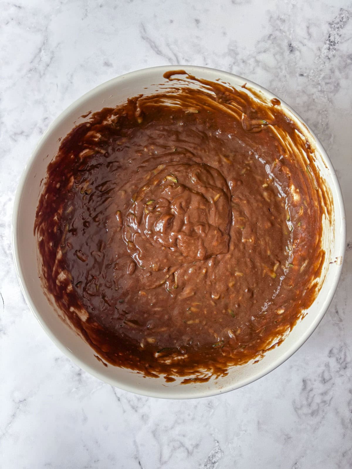 A white bowl filled with chocolate cake or brownie batter sits on a light marble countertop. The batter appears thick and slightly textured, ready to be baked.