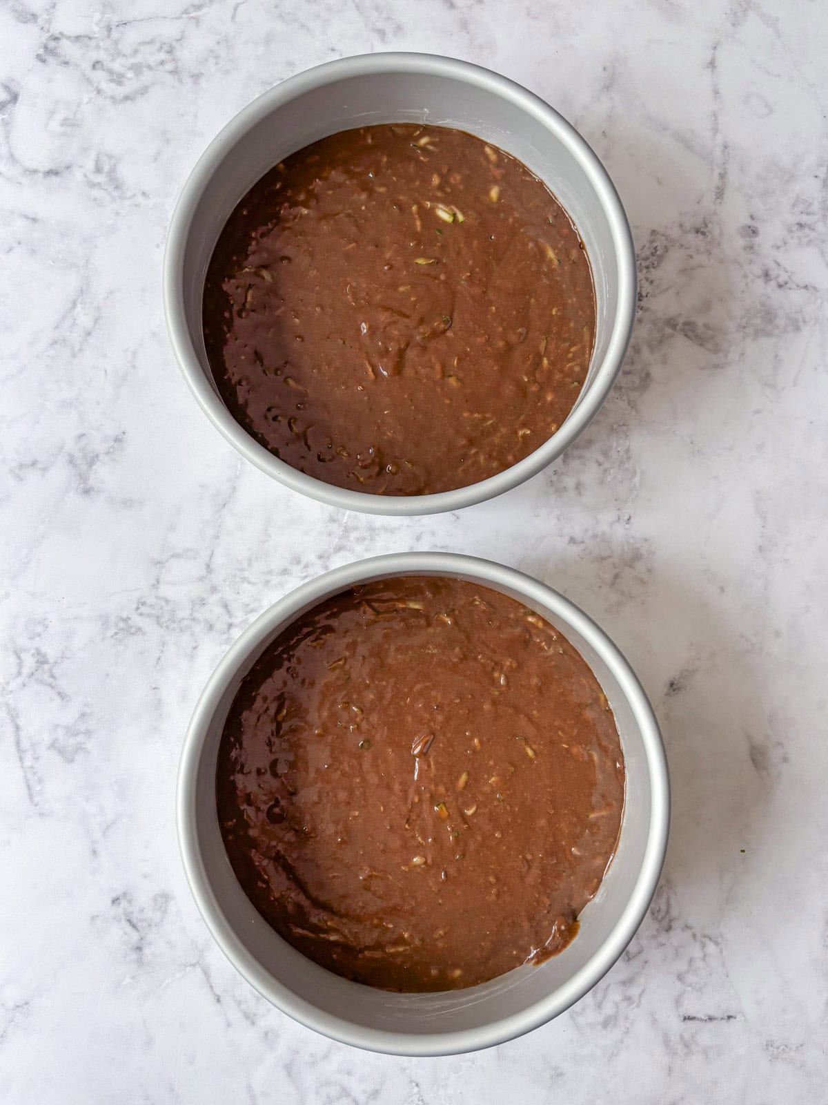 Two round cake pans filled with chocolate cake batter sit on a white marble surface, ready to be baked.