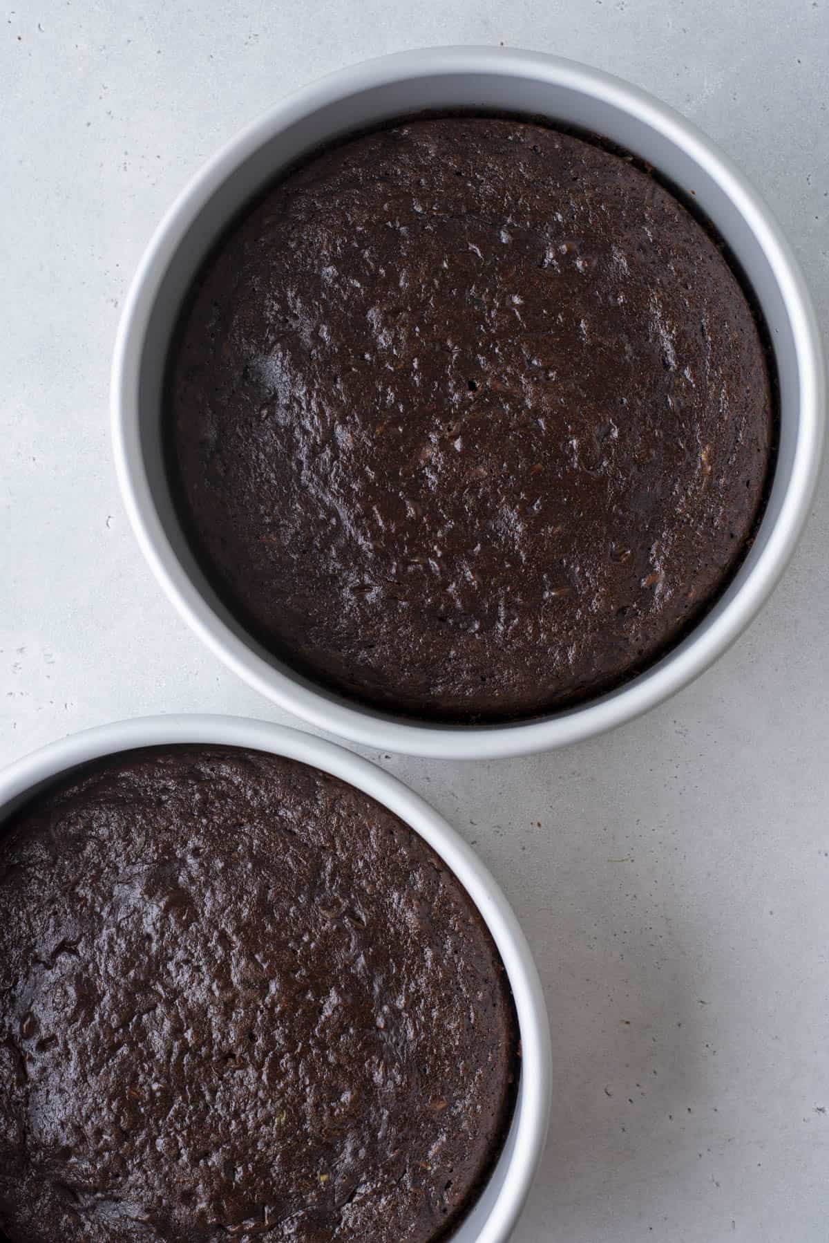 Two round cake pans filled with freshly baked chocolate cake, shown from above on a light gray surface. The cakes have a moist, slightly glossy top.