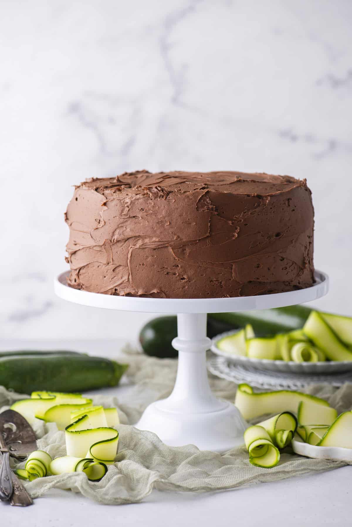 A chocolate frosted layer cake sits on a white cake stand, surrounded by green zucchini ribbons and whole zucchinis on a light surface with a marble background.