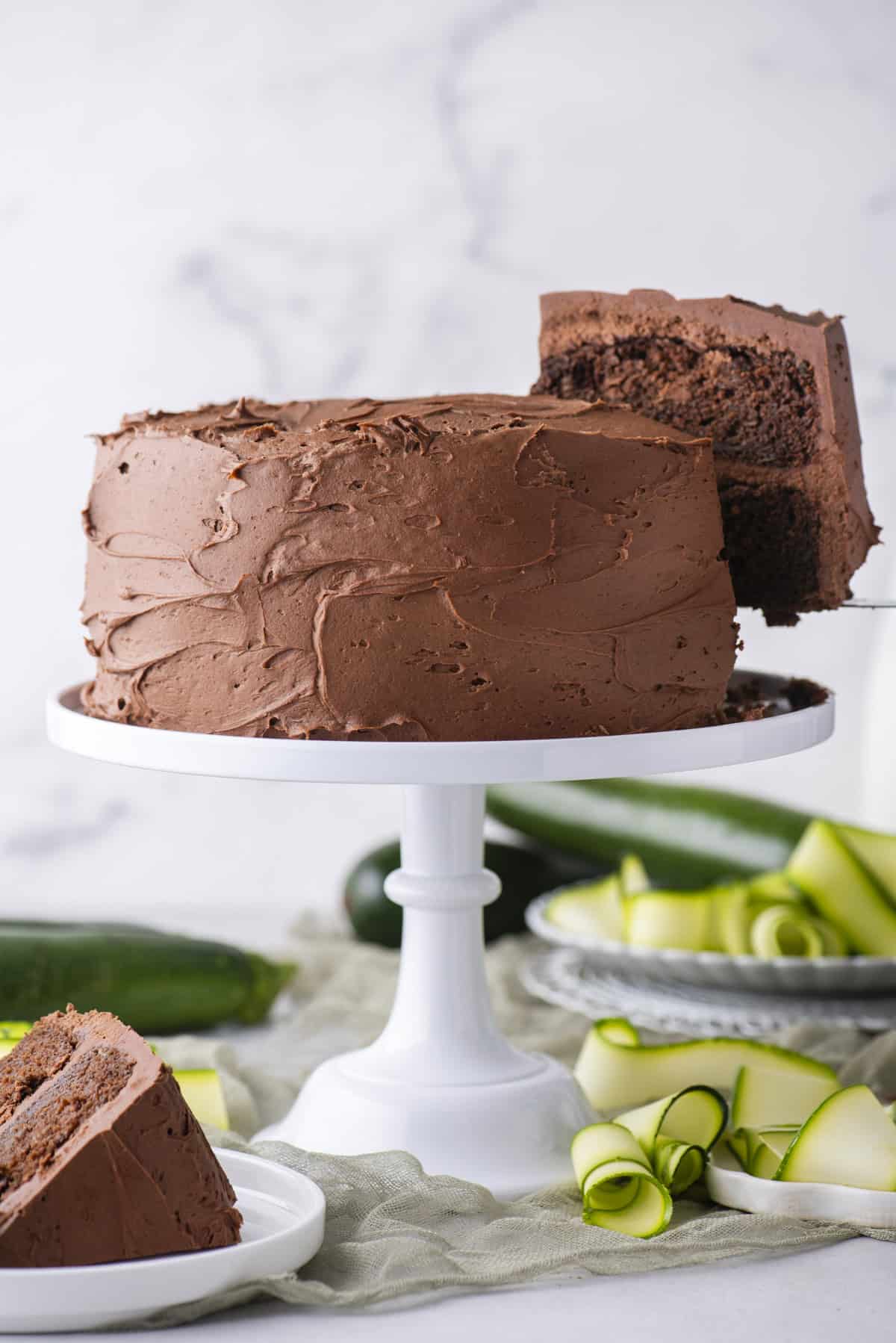 A chocolate frosted layer cake sits on a white cake stand, with one slice being served. Zucchini ribbons and a plate with another cake slice are nearby on a light-colored surface.
