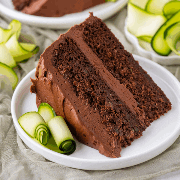 A slice of chocolate cake with chocolate frosting is served on a white plate, garnished with thin, rolled slices of zucchini. More zucchini rolls and a second plate are visible in the background.