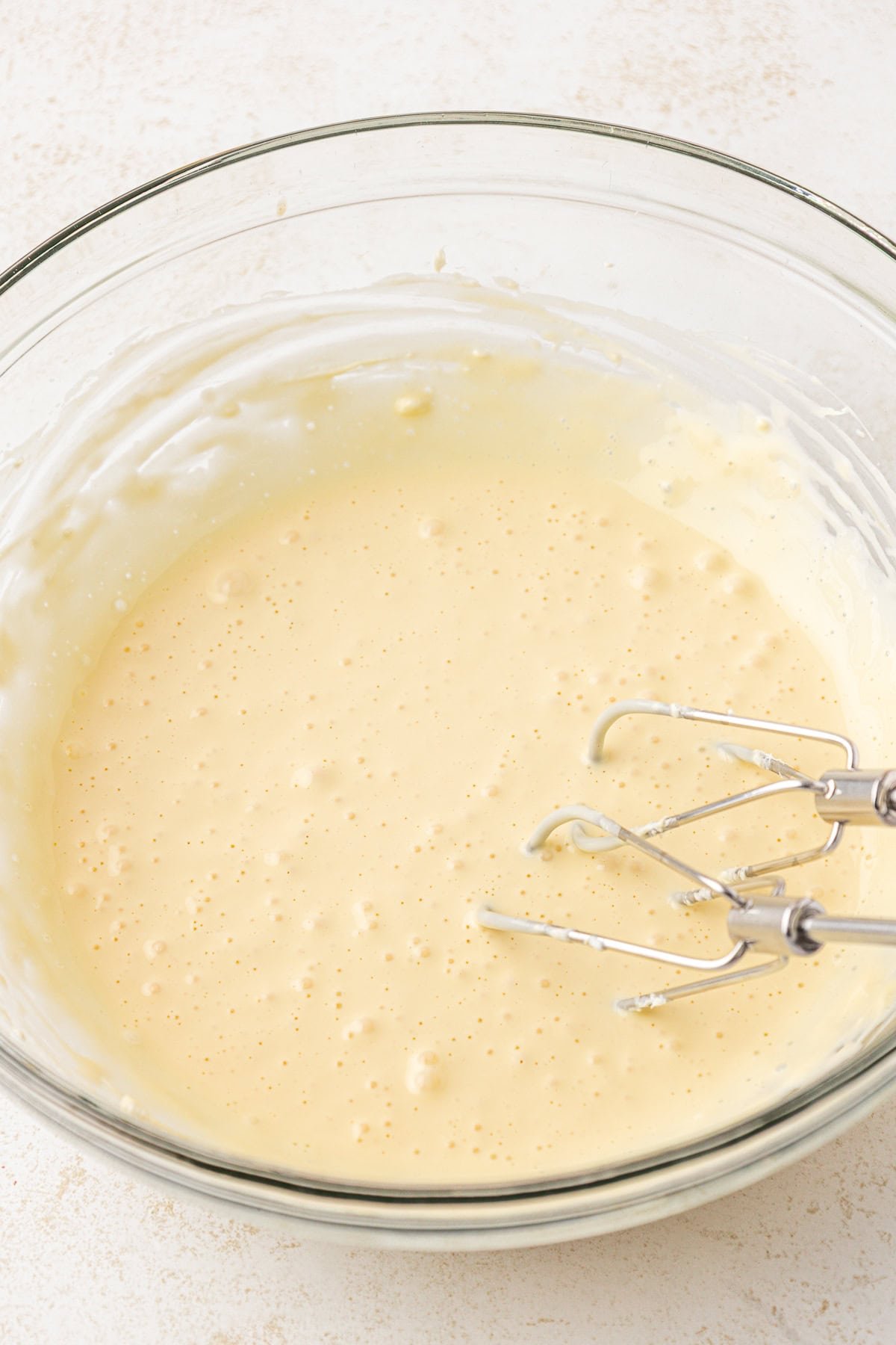 A glass mixing bowl filled with pale yellow cake batter sits on a light surface. An electric hand mixer with beaters is resting in the bowl, partially coated with batter.