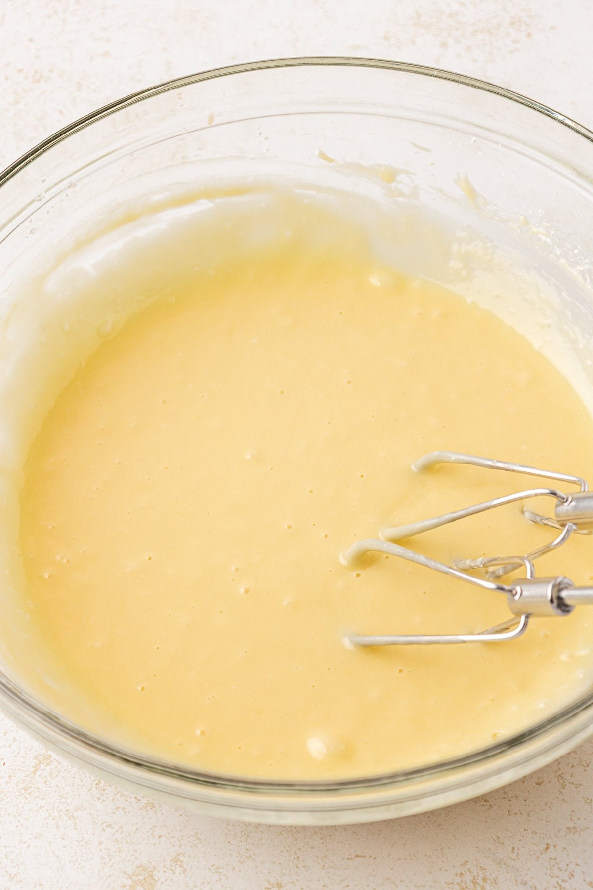 A glass mixing bowl filled with smooth yellow cake batter. Metal beaters from a hand mixer are resting in the bowl, partially coated with batter. The bowl sits on a light-colored surface.