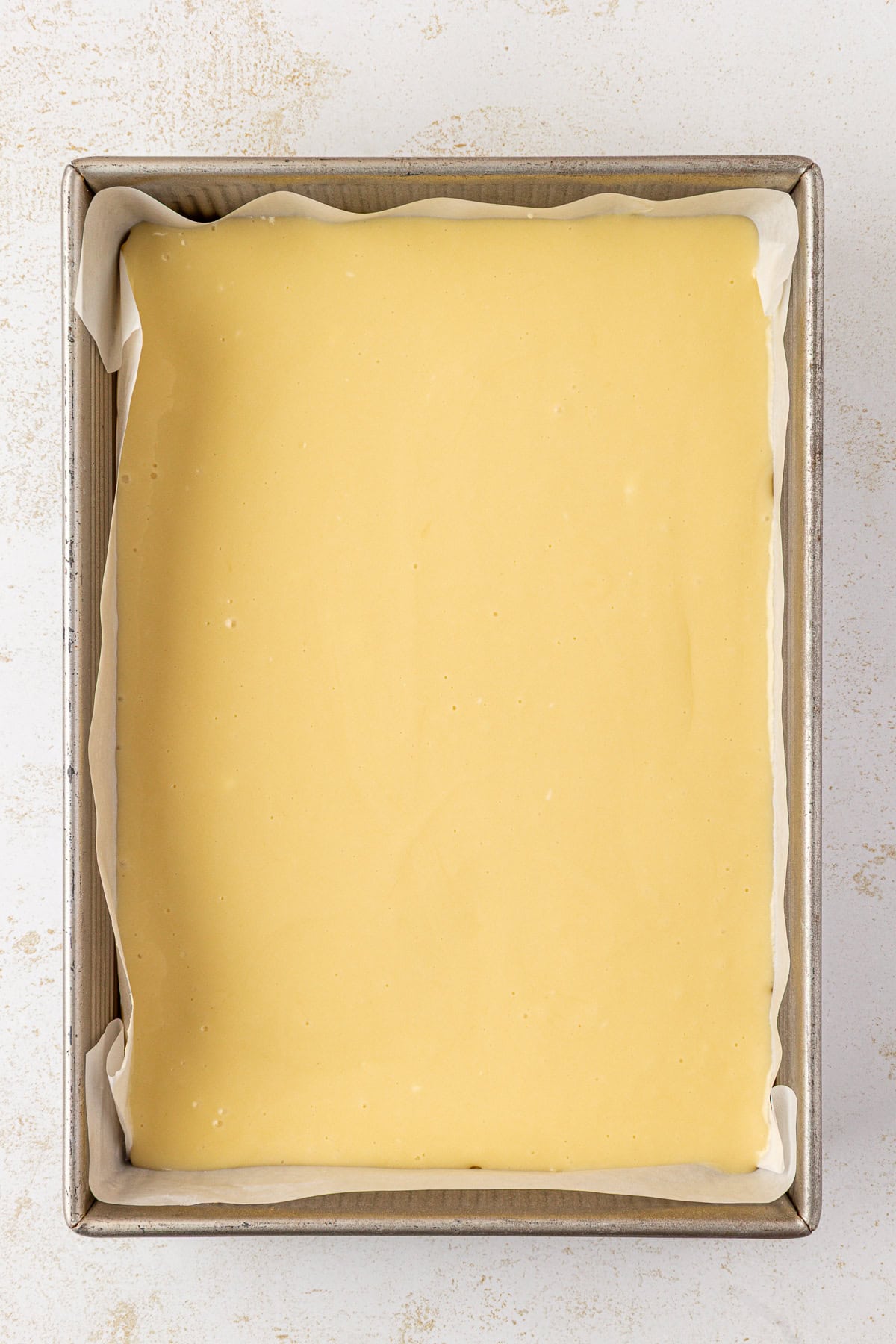 A square baking pan lined with parchment paper filled with smooth, uncooked yellow batter, ready to be baked. The pan is set on a light-colored surface.
