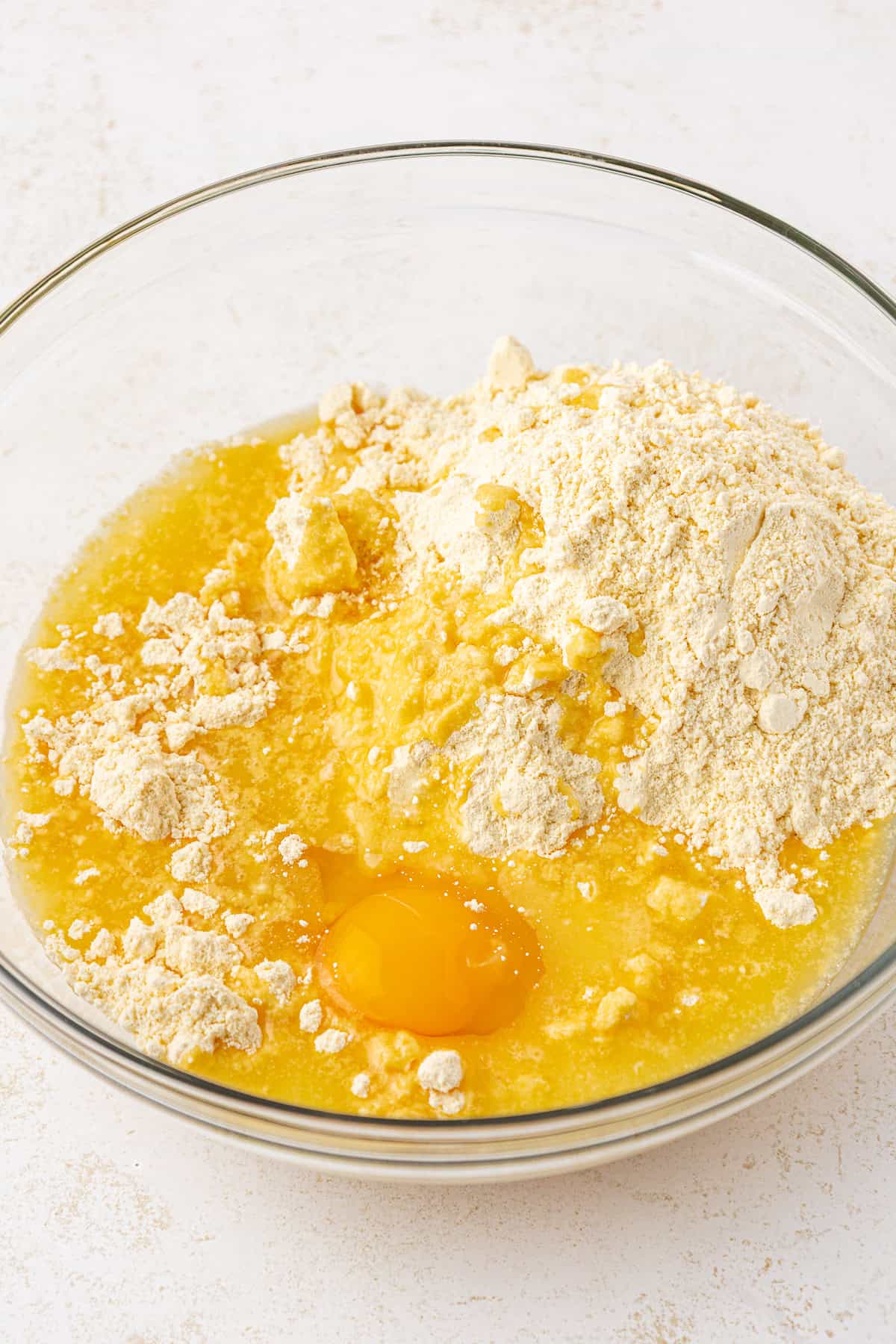A clear glass bowl filled with flour, a cracked raw egg, and melted butter on a light-colored surface, ready to be mixed for baking.