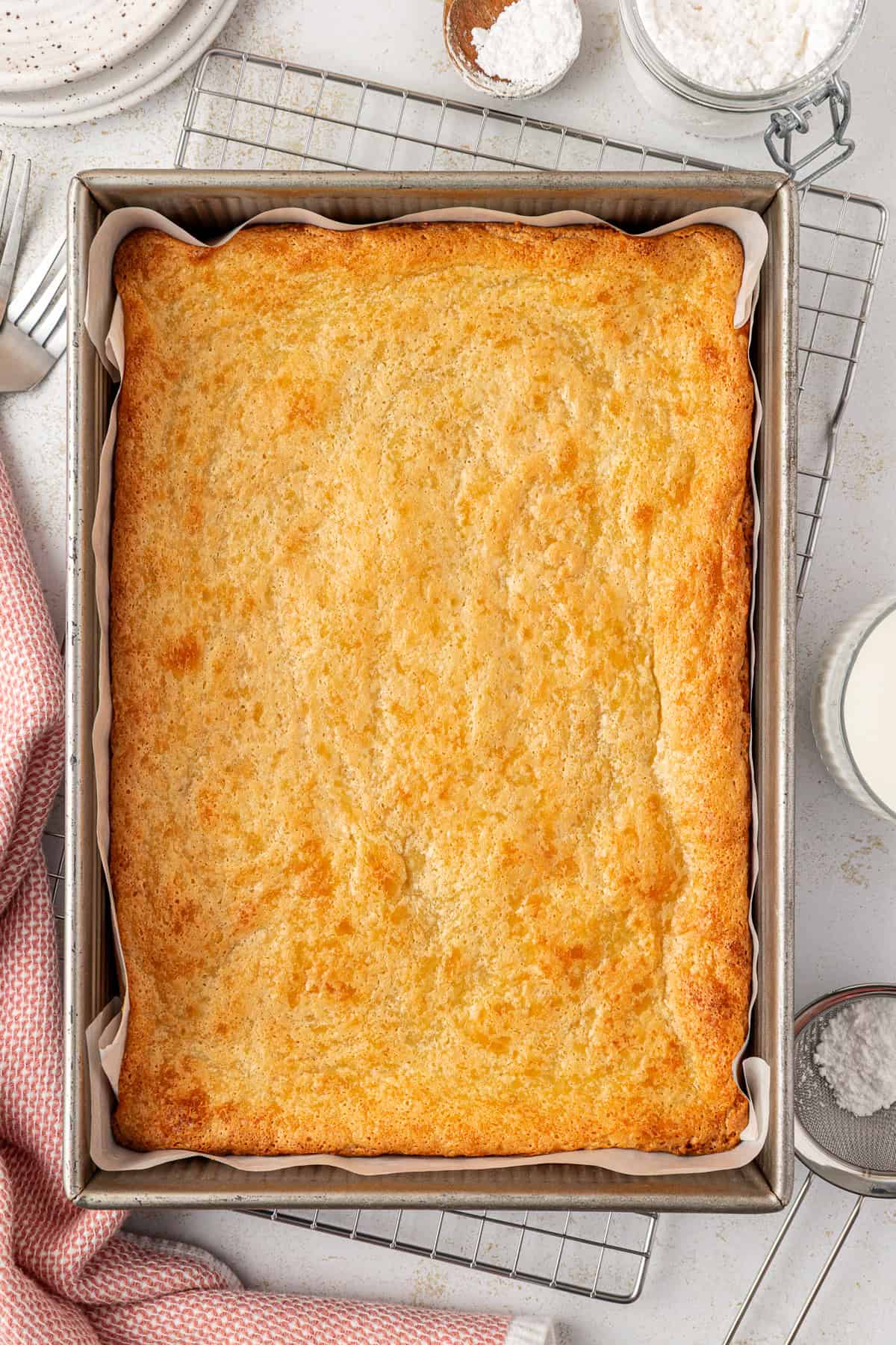A golden-brown sheet cake in a parchment-lined square baking pan sits on a cooling rack, with a pink towel, a fork, powdered sugar, and a glass of milk nearby.