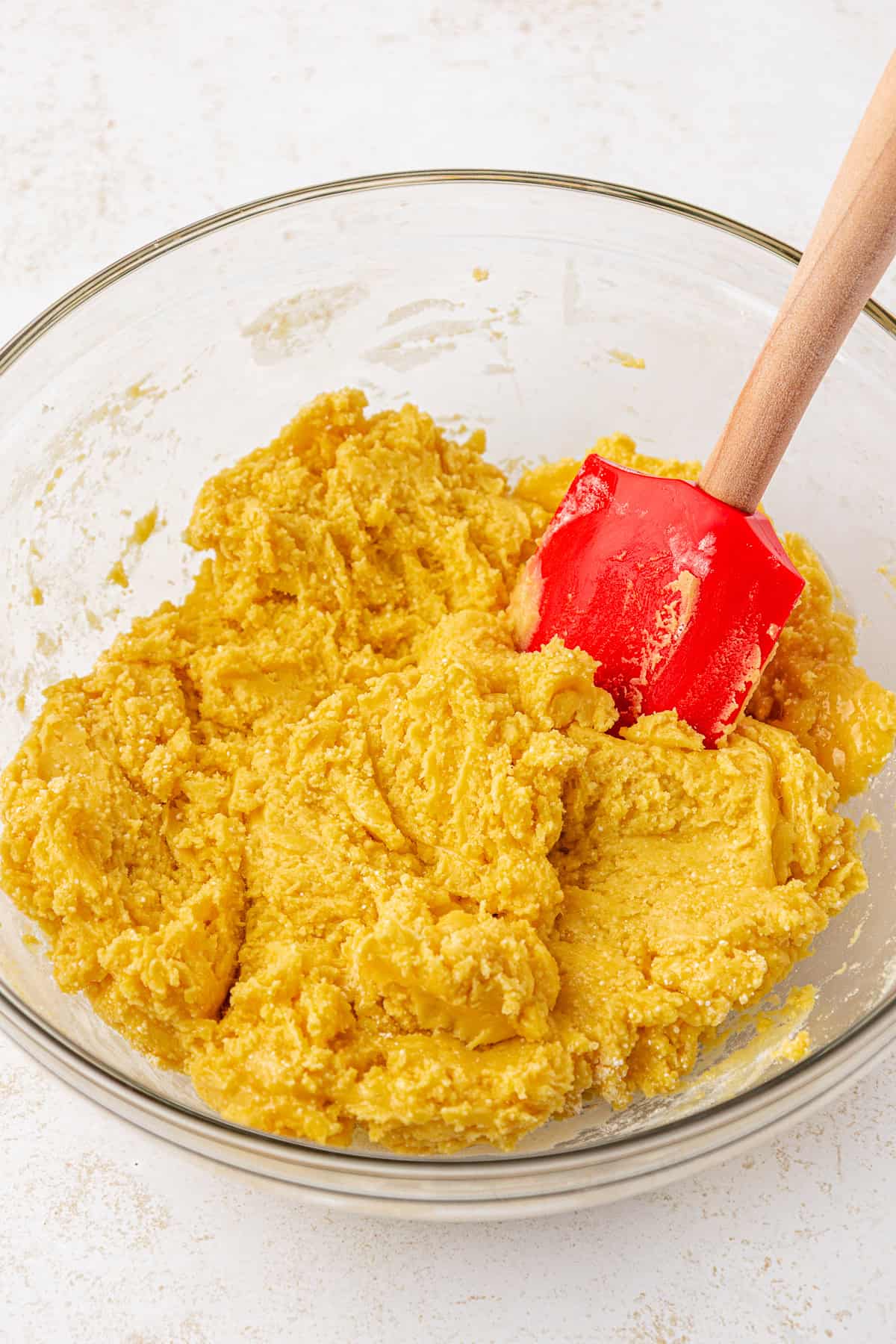 A glass bowl filled with yellow cookie dough being mixed with a red silicone spatula on a light countertop.