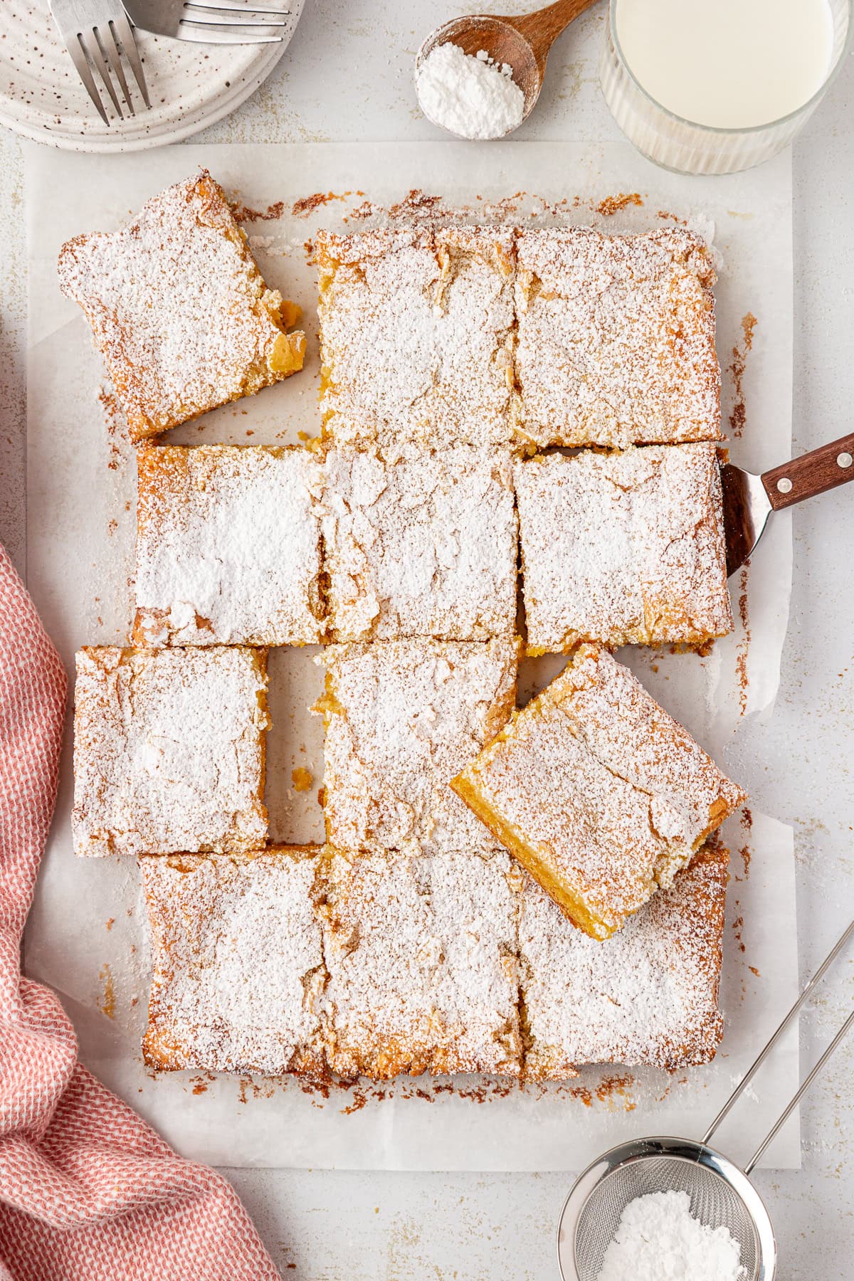 A tray of lemon bars cut into squares, topped generously with powdered sugar. One bar is being lifted with a spatula; a pink kitchen towel, a glass of milk, and utensils are nearby on a white surface.
