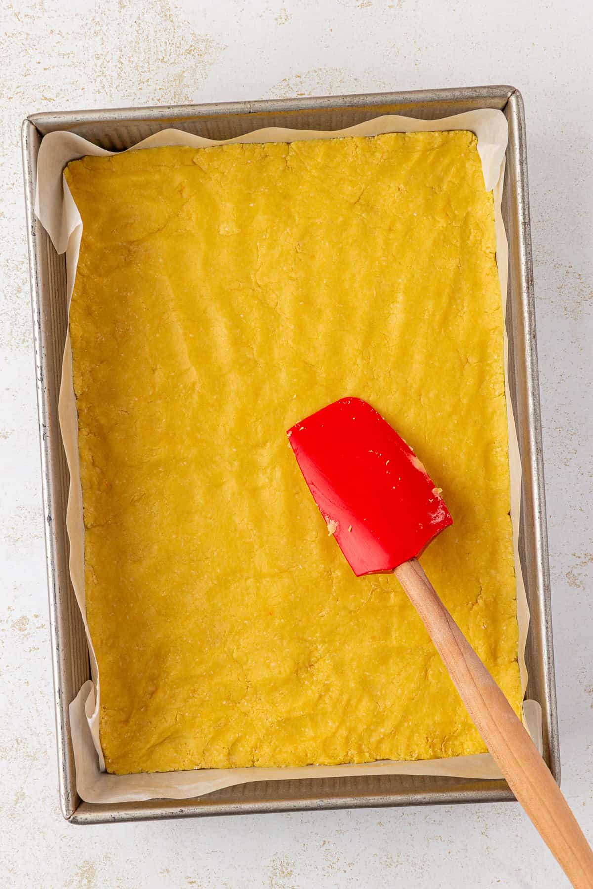 A rectangular baking pan lined with parchment paper holds an even layer of yellow dough being spread by a red silicone spatula with a wooden handle.