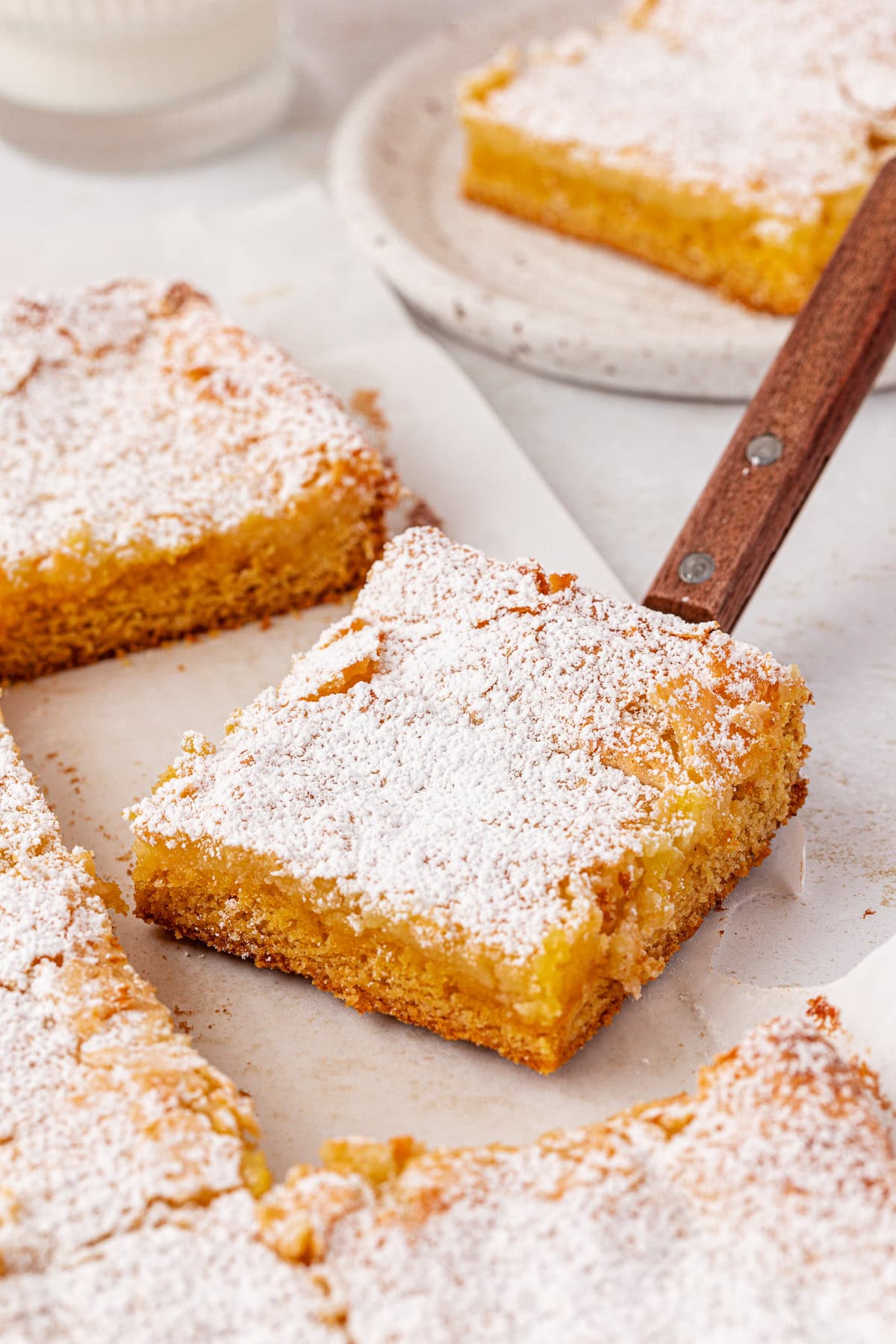 A close-up of several square lemon bars topped with powdered sugar, arranged on parchment paper with one bar lifted slightly by a spatula.