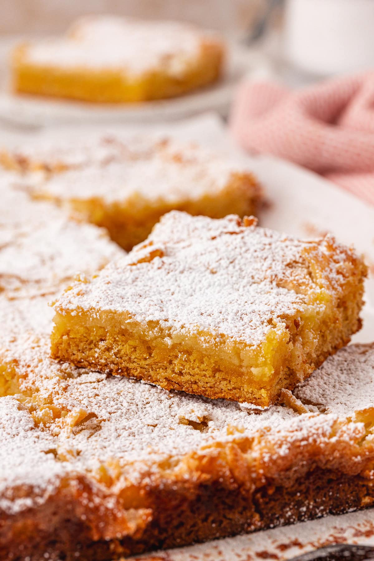 A close-up of lemon bars topped with powdered sugar, with one bar slightly raised above the rest, showing a golden, moist texture and crumbly edges. A pink cloth is blurred in the background.