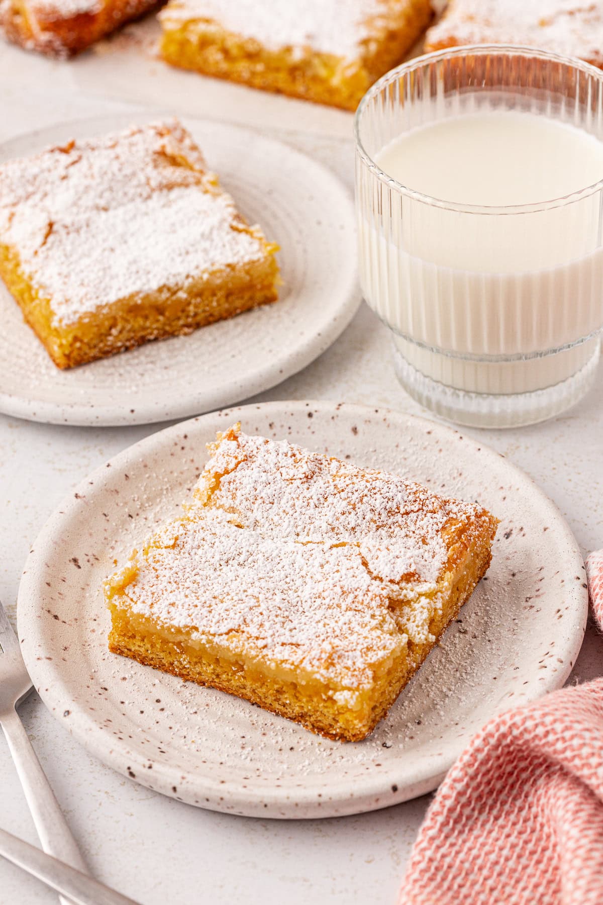 A plate with two powdered sugar-dusted lemon bars sits beside a glass of milk; another plate with a single lemon bar is visible in the background, along with a pink textured napkin.