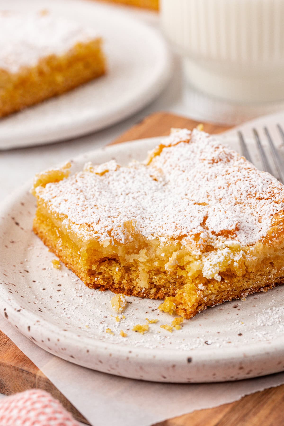 A close-up of a square piece of gooey butter cake dusted with powdered sugar on a speckled plate, with a fork, another slice, and a glass of milk in the background.