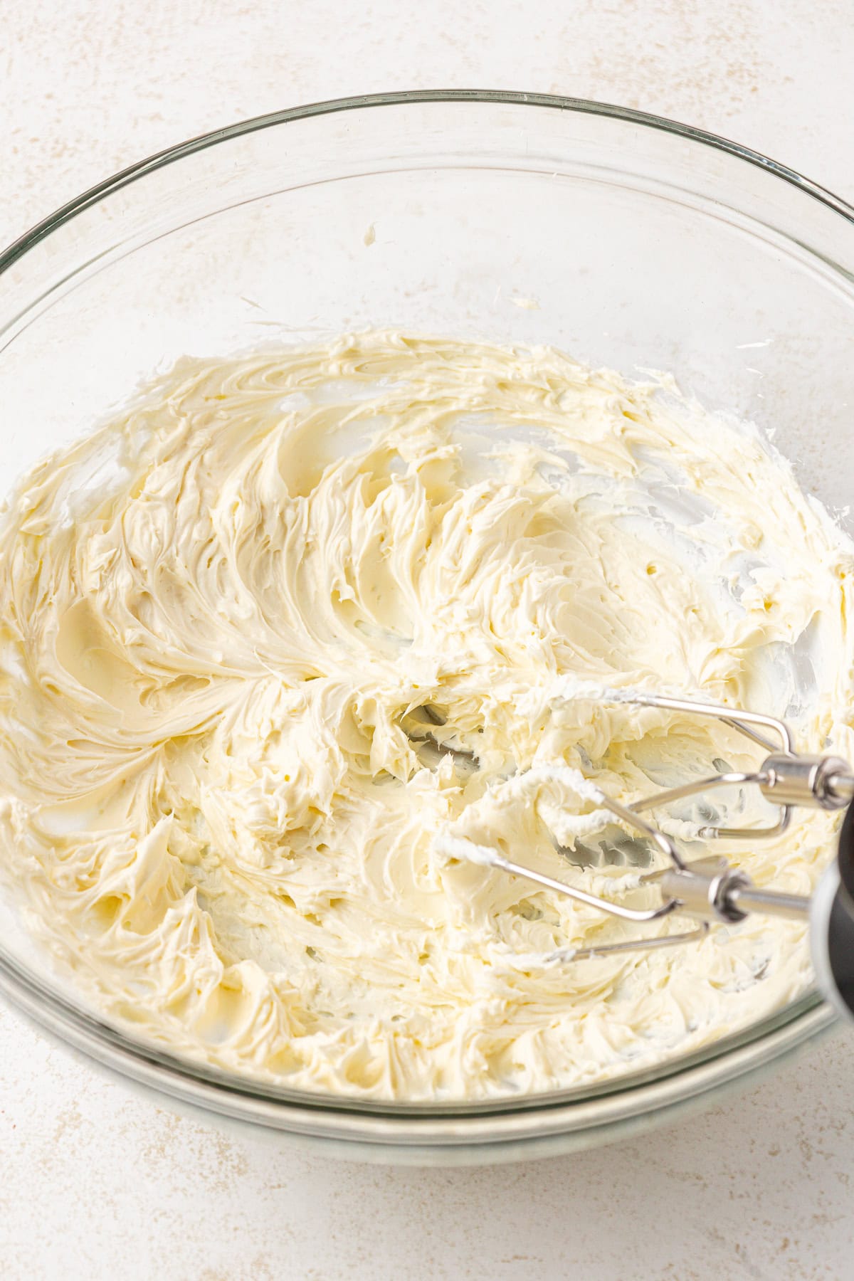 A hand mixer with beaters rests in a glass bowl filled with creamy, whipped butter mixture, showing smooth and fluffy texture on a light, speckled surface.