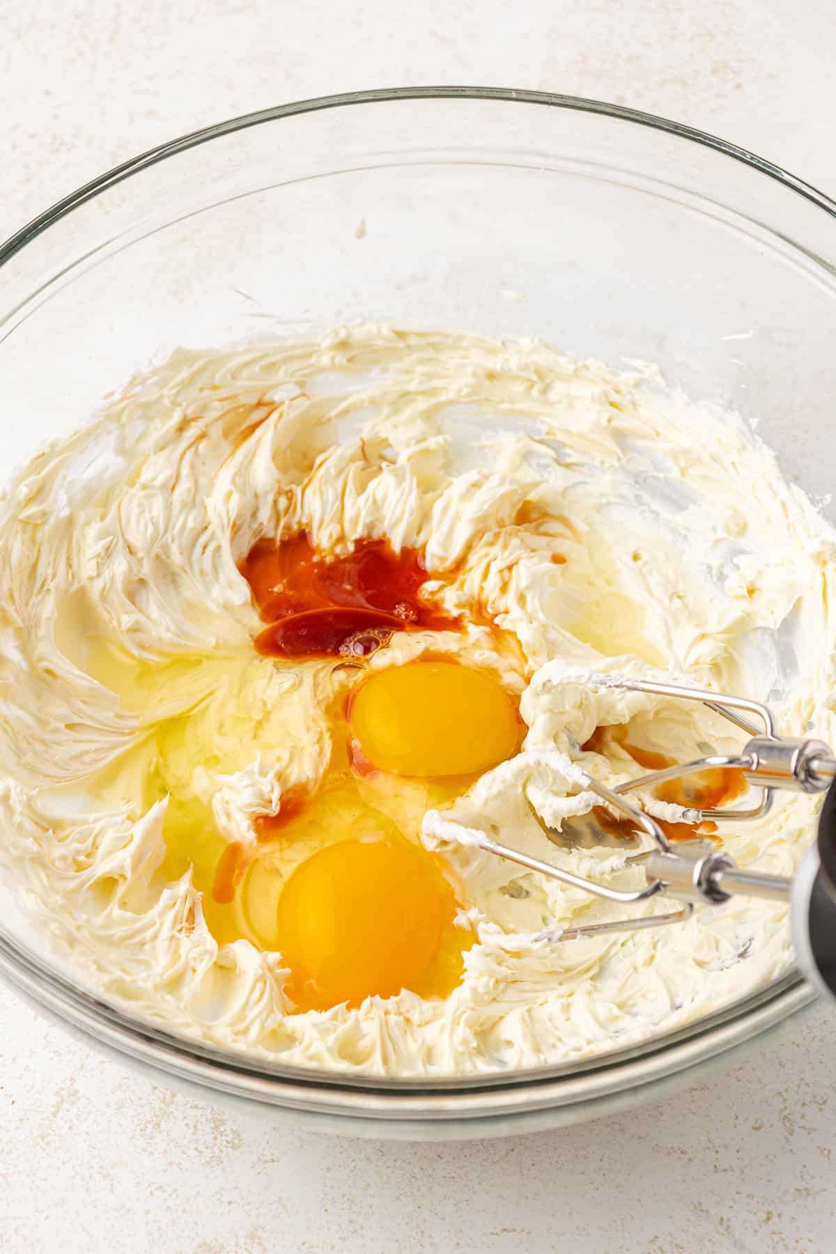 A glass bowl contains whipped cream cheese, two raw eggs, vanilla extract, and sugar being mixed with an electric hand mixer on a light countertop.
