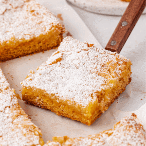 A square piece of golden, crumbly cake dusted with powdered sugar is being lifted from a tray with a metal spatula. Other pieces of cake are visible on parchment paper in the background.