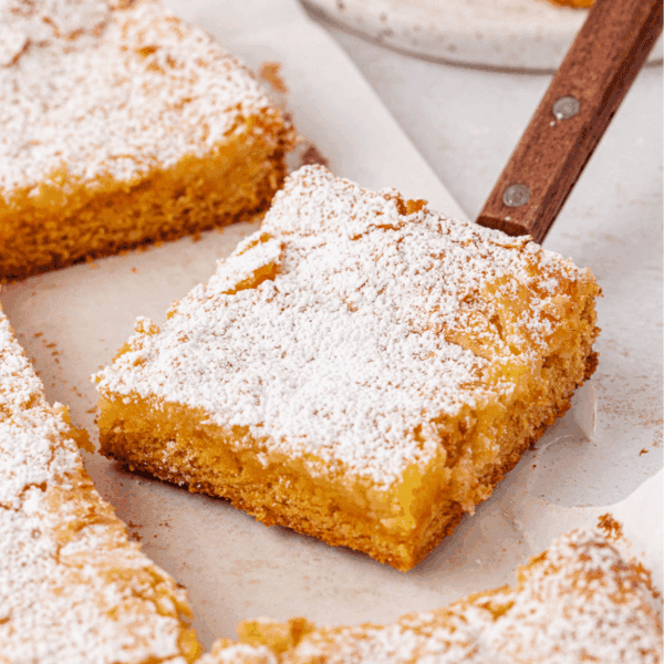 A square piece of golden, crumbly cake dusted with powdered sugar is being lifted from a tray with a metal spatula. Other pieces of cake are visible on parchment paper in the background.
