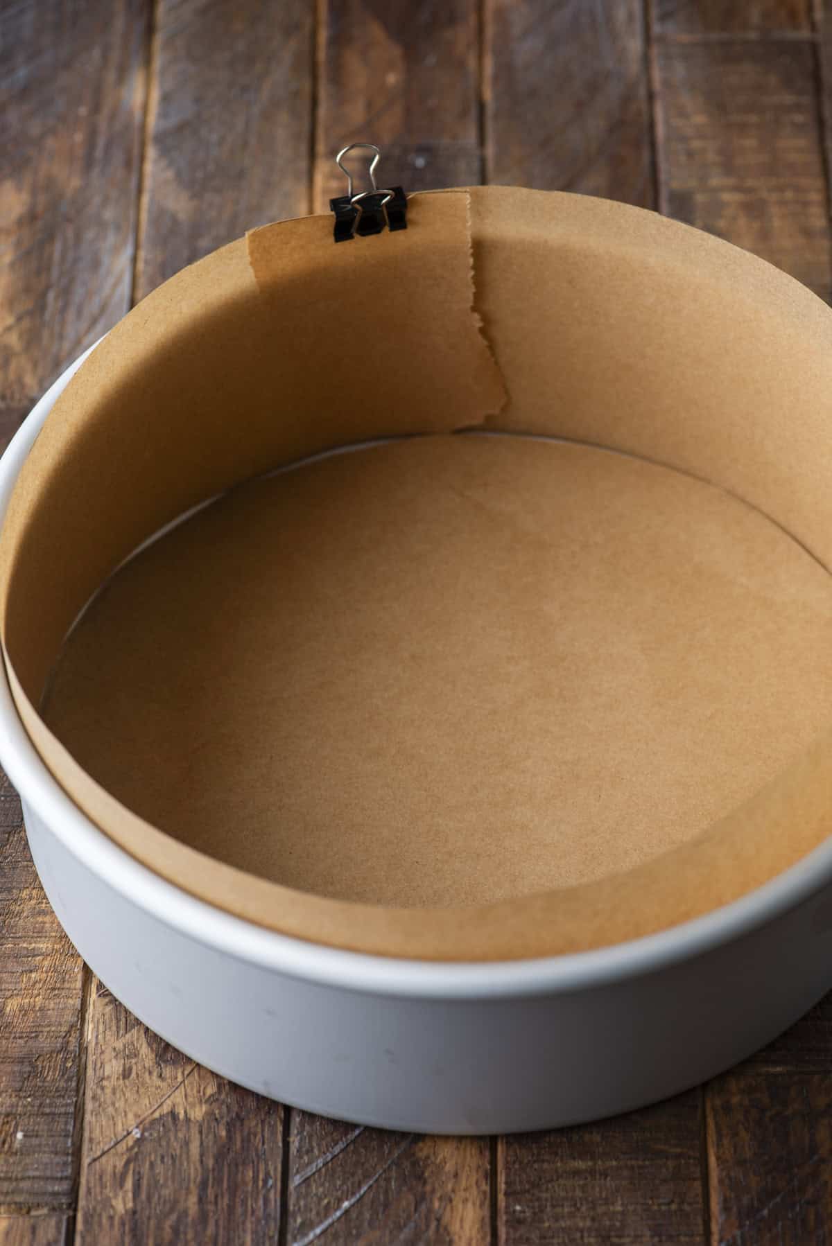 A round cake pan lined with brown parchment paper along the bottom and sides, held in place with a black binder clip, sits on a rustic wooden surface.