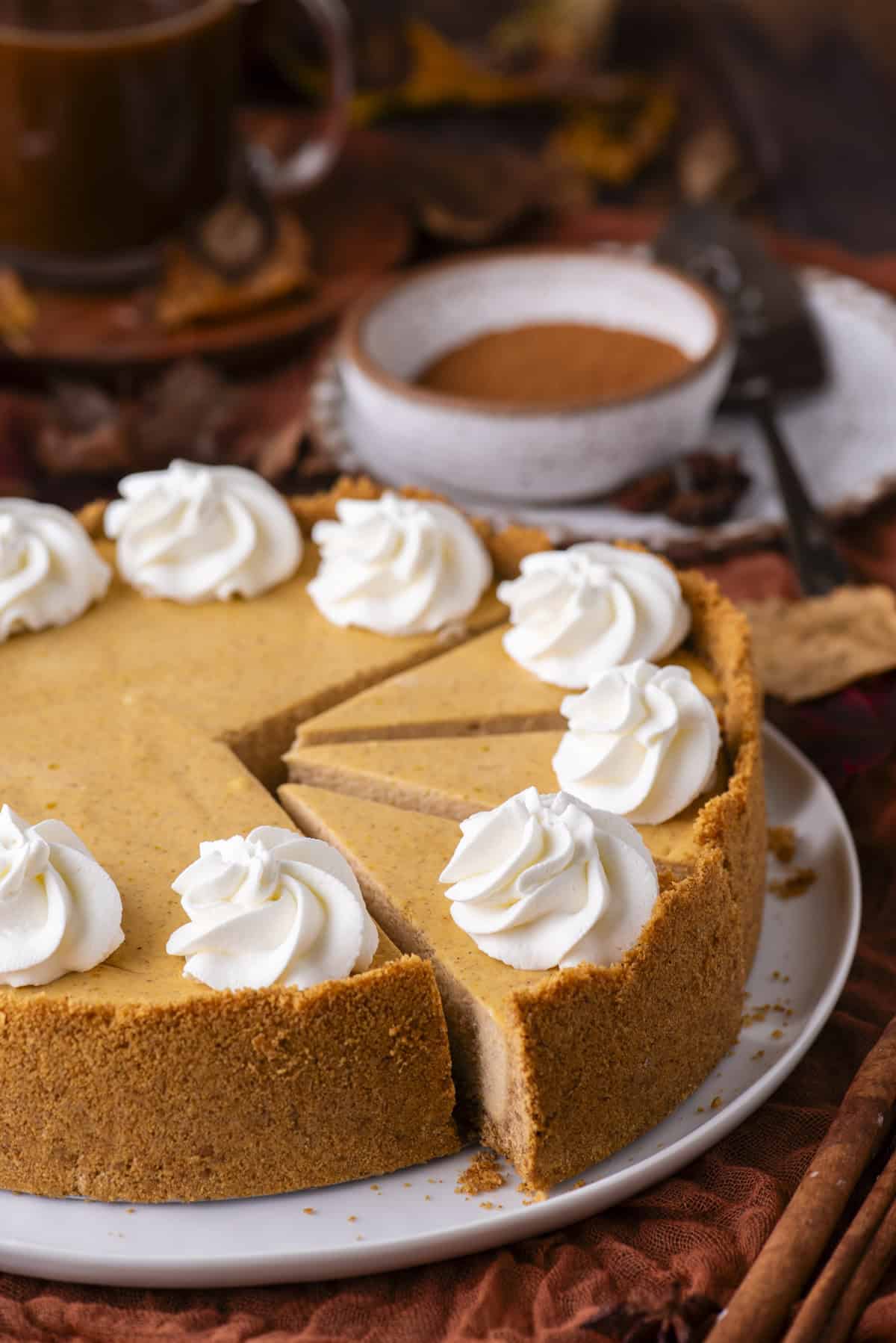 A pumpkin cheesecake with a graham cracker crust, topped with dollops of whipped cream, sits on a plate with a slice removed. Nearby are a bowl of cinnamon, a cup of coffee, and autumn leaves.