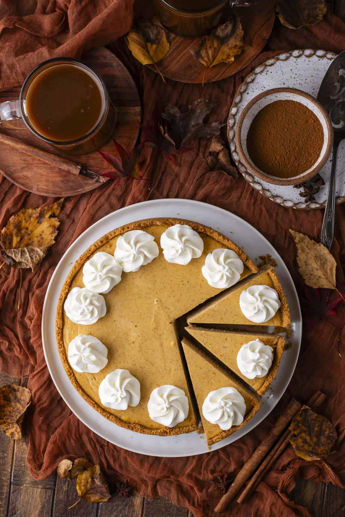 A whole pumpkin pie with whipped cream dollops on top, two slices cut, surrounded by a cup of coffee, bowl of cinnamon, fall leaves, and a rust-colored cloth on a wooden table.