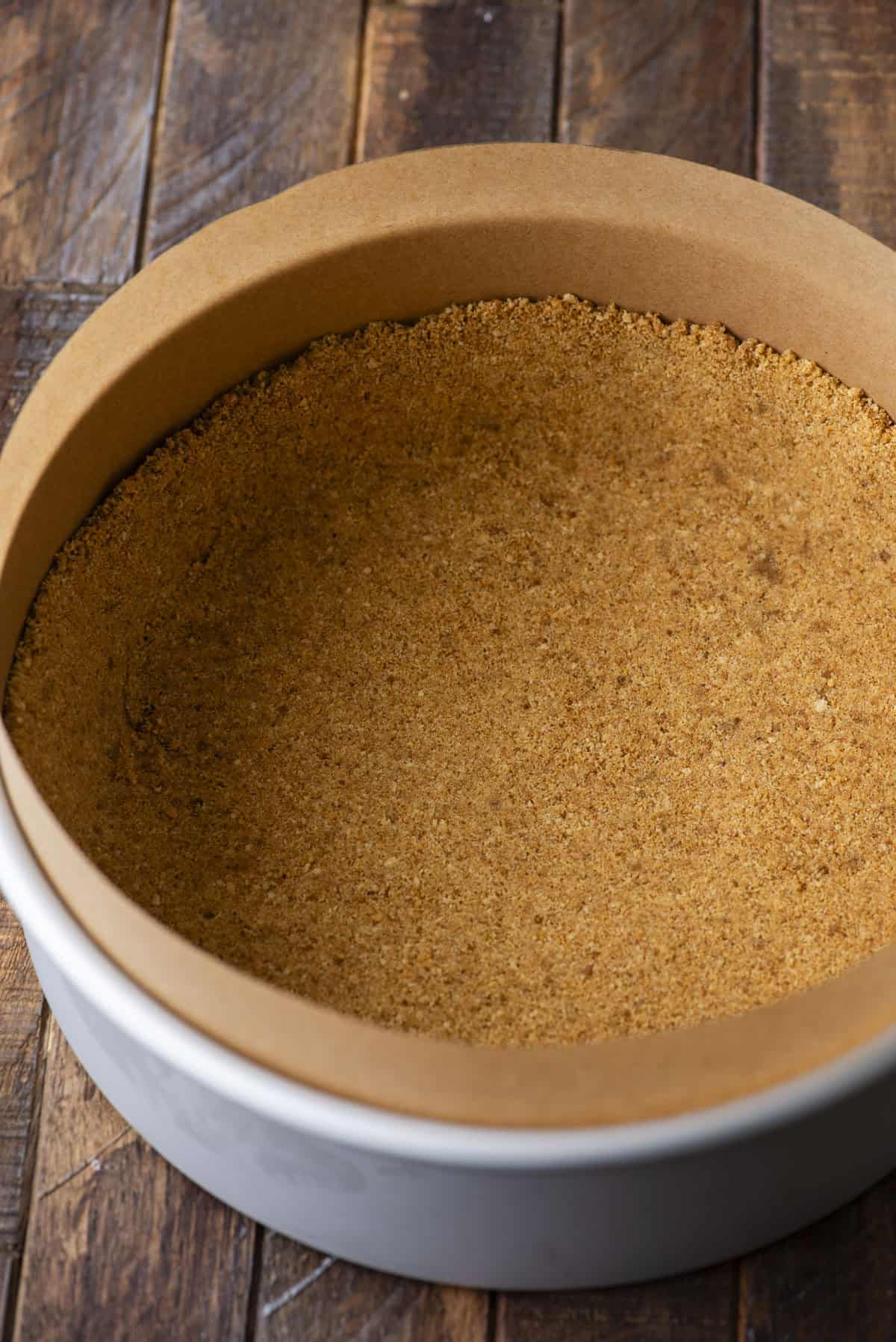A round baking pan lined with parchment paper and filled with a pressed graham cracker crust, set on a rustic wooden surface.