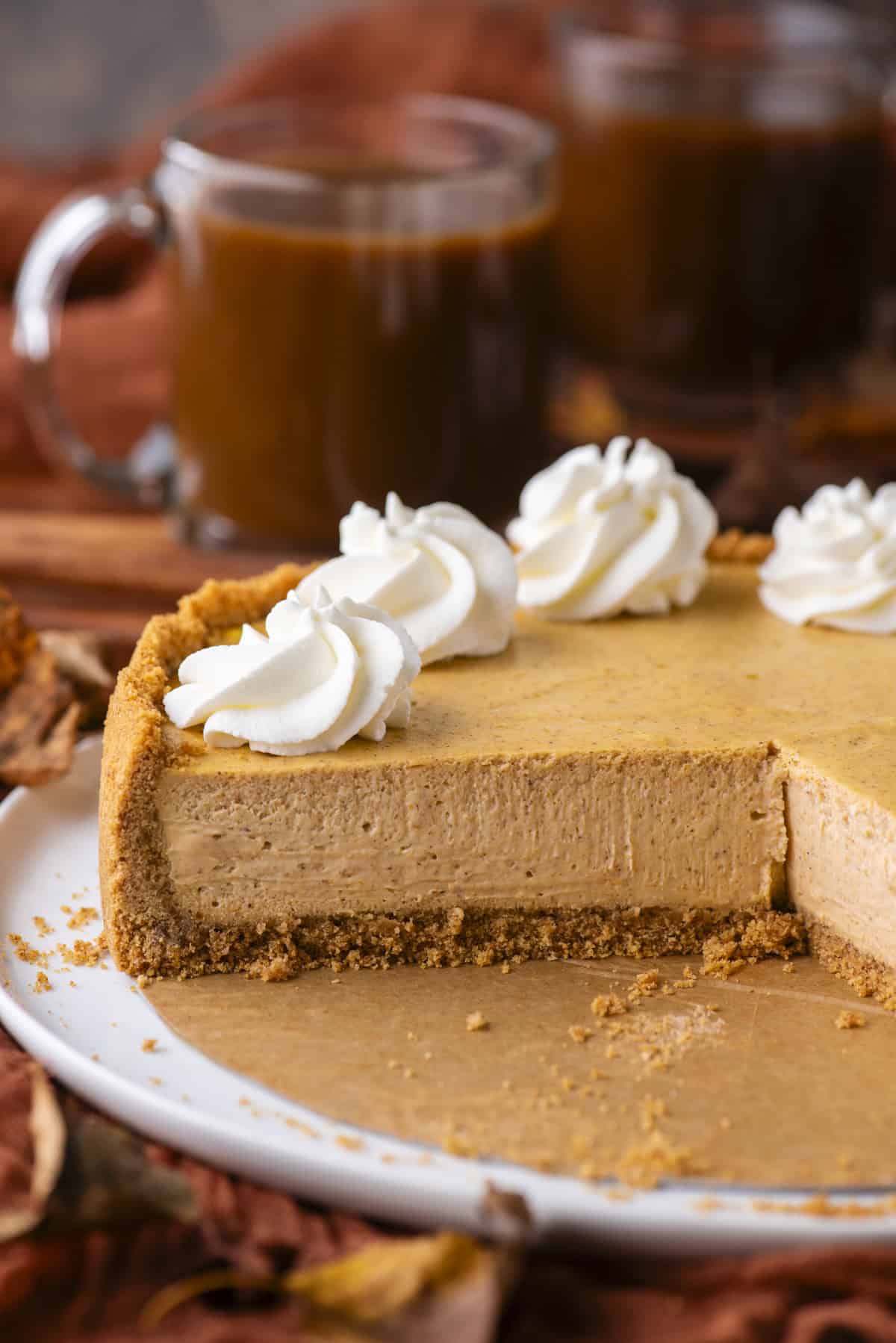 A pumpkin cheesecake with a graham cracker crust sits on a white plate. A slice has been removed, revealing a creamy filling. The cheesecake is topped with whipped cream swirls, and coffee mugs are in the background.