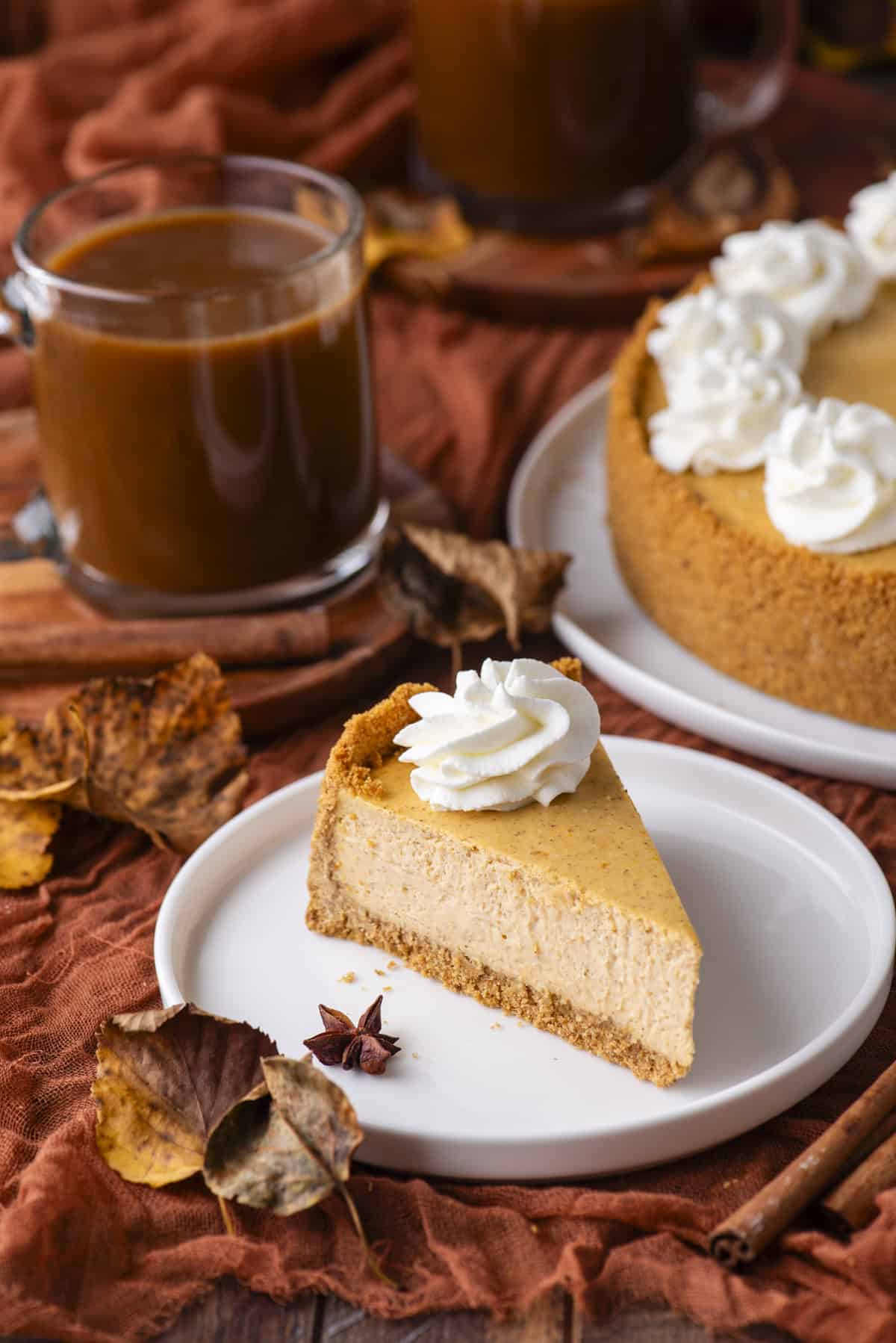 A slice of pumpkin cheesecake topped with whipped cream on a white plate, surrounded by autumn leaves, spices, and a glass mug of coffee. The whole cheesecake and another mug are visible in the background.