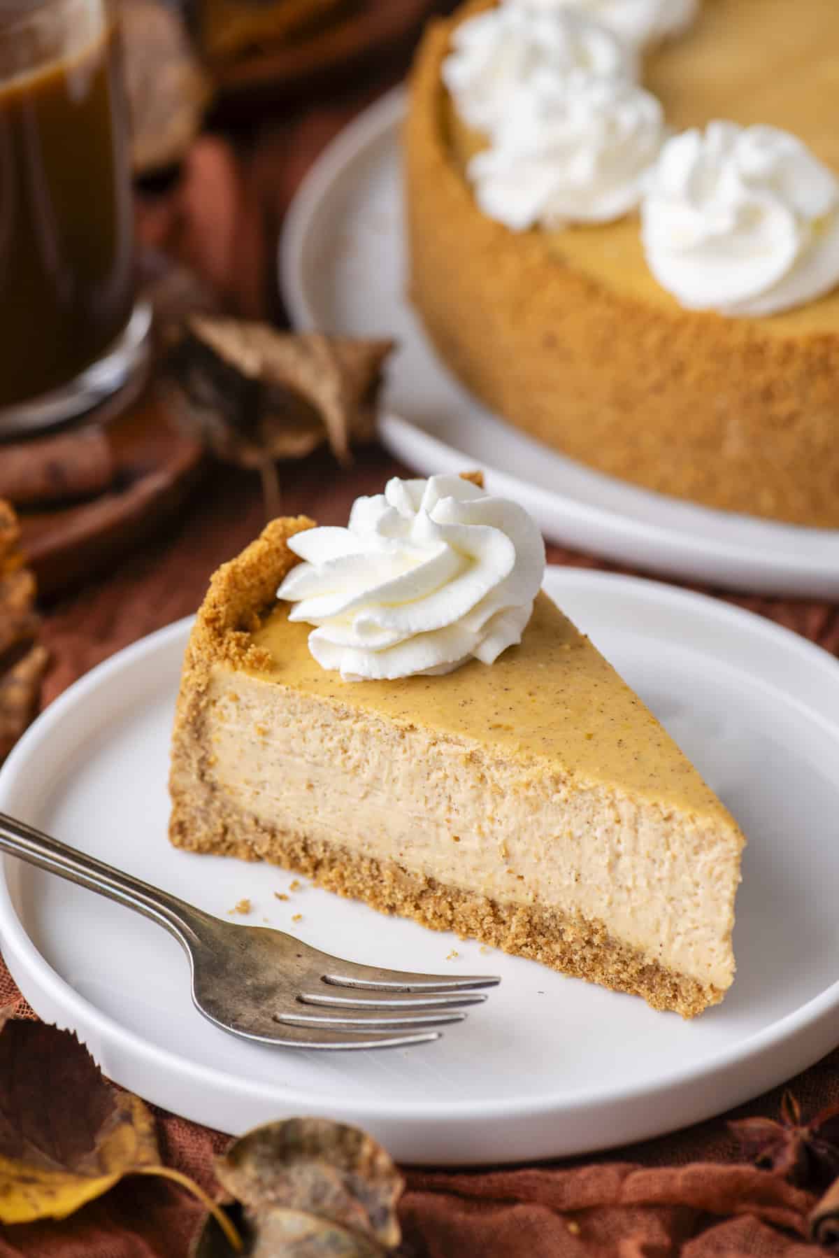 A slice of pumpkin cheesecake with a graham cracker crust and a swirl of whipped cream on top, served on a white plate with a fork. The whole cheesecake and autumn leaves are in the background.