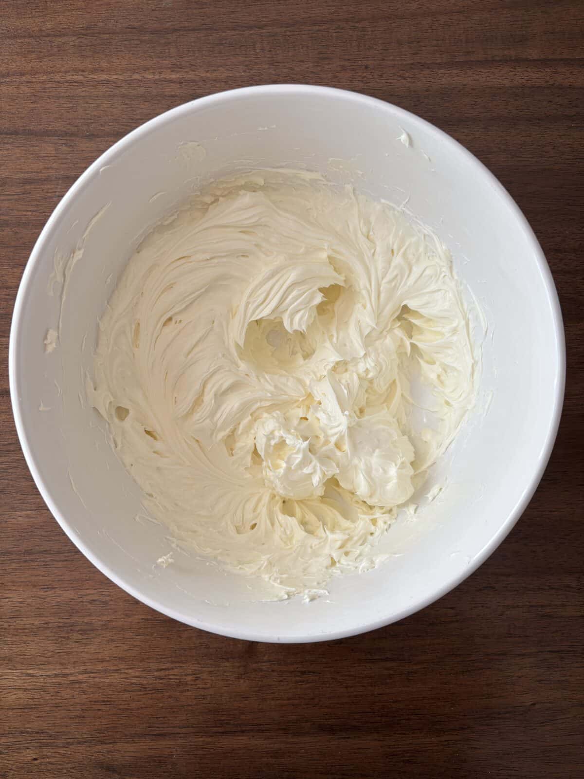 A large white mixing bowl filled with creamy, whipped frosting or batter sits on a dark wooden surface. The frosting has a smooth and fluffy texture with visible swirls from mixing.