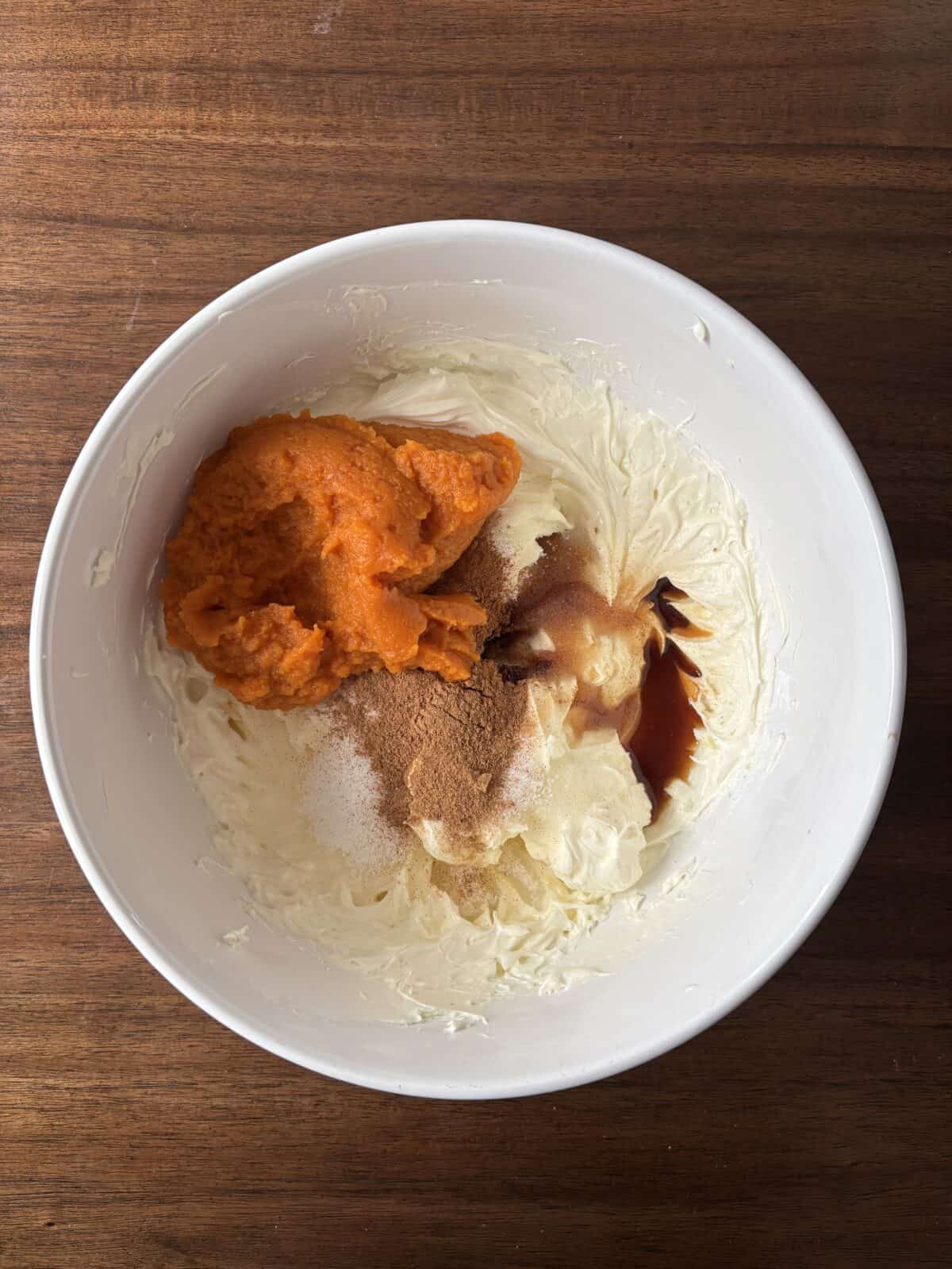 A white bowl on a wooden surface contains cream cheese, pumpkin puree, brown sugar, vanilla extract, and spices, ready to be mixed together.