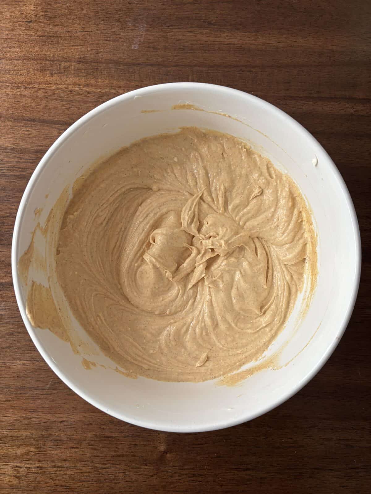 A white mixing bowl containing a light brown, creamy batter sits on a wooden surface. The batter has visible swirls from mixing.