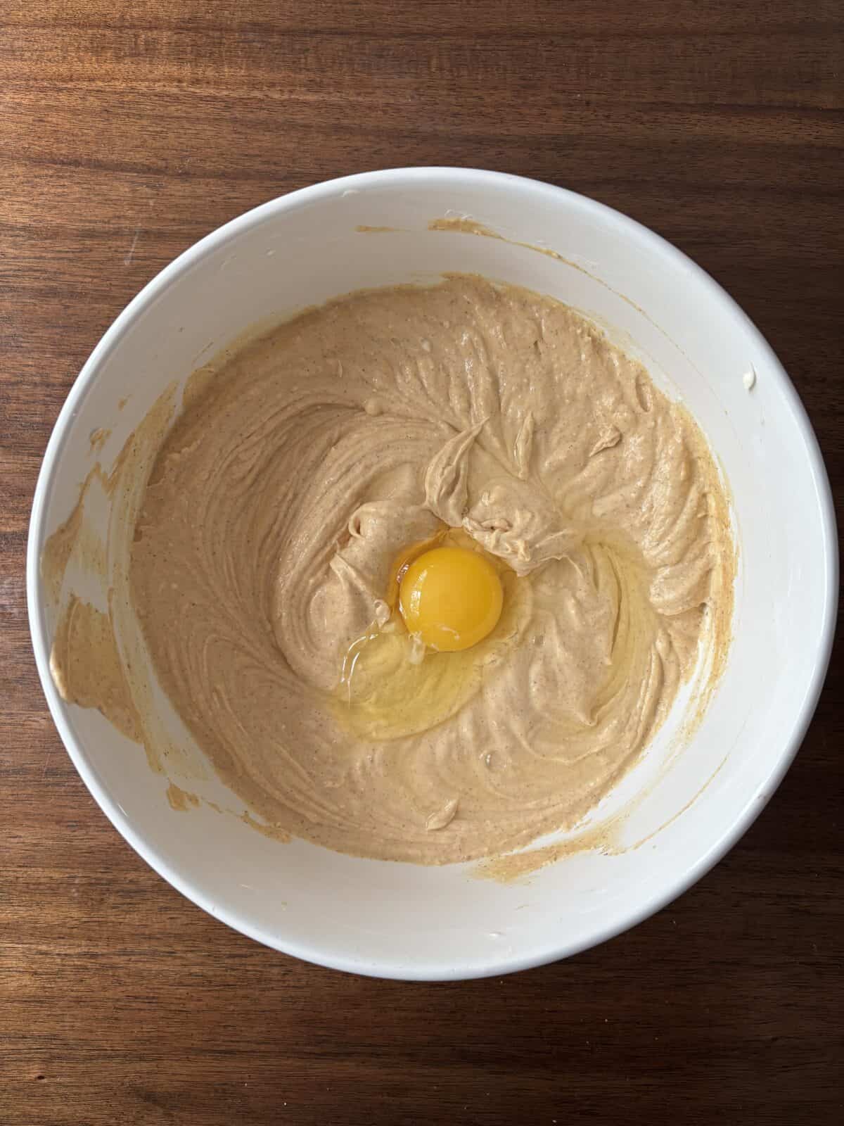 A white mixing bowl with creamy light brown batter and a raw egg on top sits on a dark wooden surface.