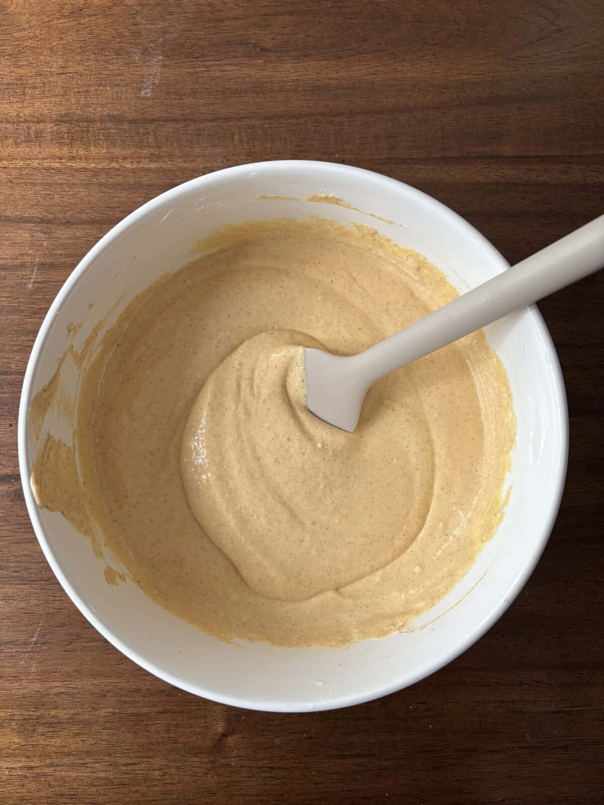 A white mixing bowl filled with light brown batter and a white spatula resting inside, placed on a wooden surface.