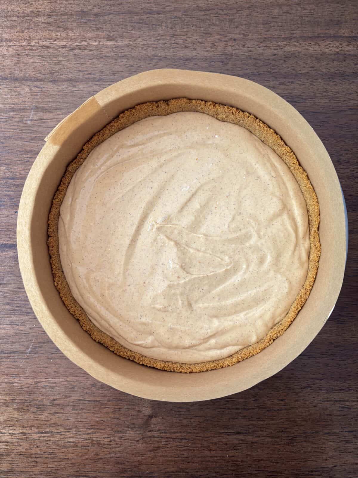 A round pie with a graham cracker crust and a smooth, creamy tan filling sits in a parchment-lined baking tin on a wooden surface.
