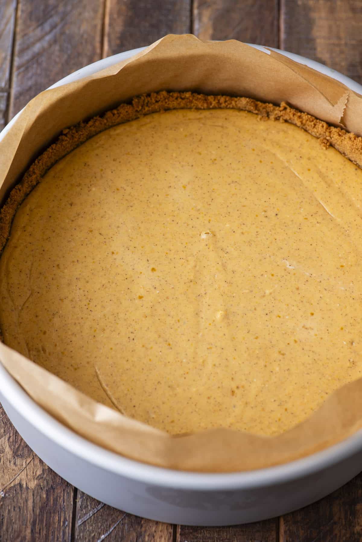 A round cheesecake with a graham cracker crust, lined with parchment paper, sits in a baking pan on a wooden surface. The filling is smooth and lightly golden, suggesting a pumpkin or spiced flavor.