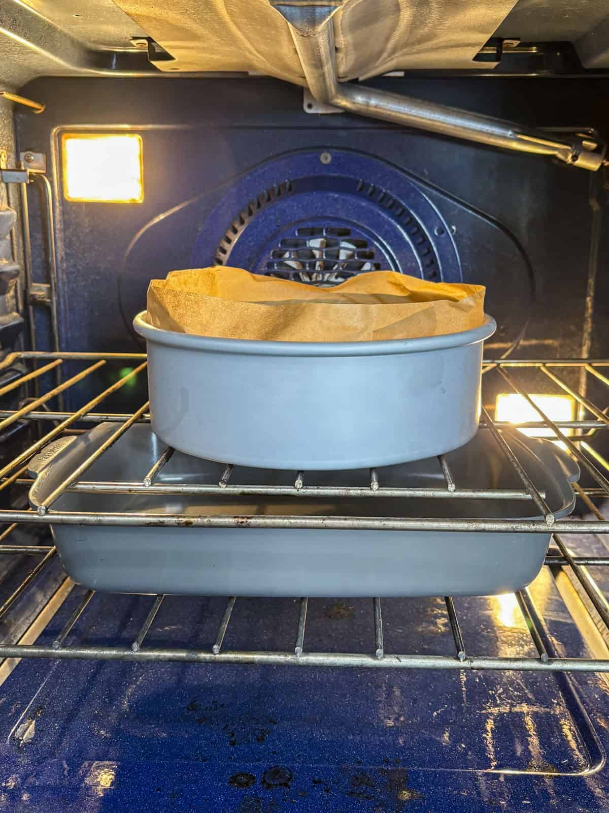 A round cake pan lined with parchment paper sits on a rectangular baking dish in an oven, with the oven light on and racks visible.
