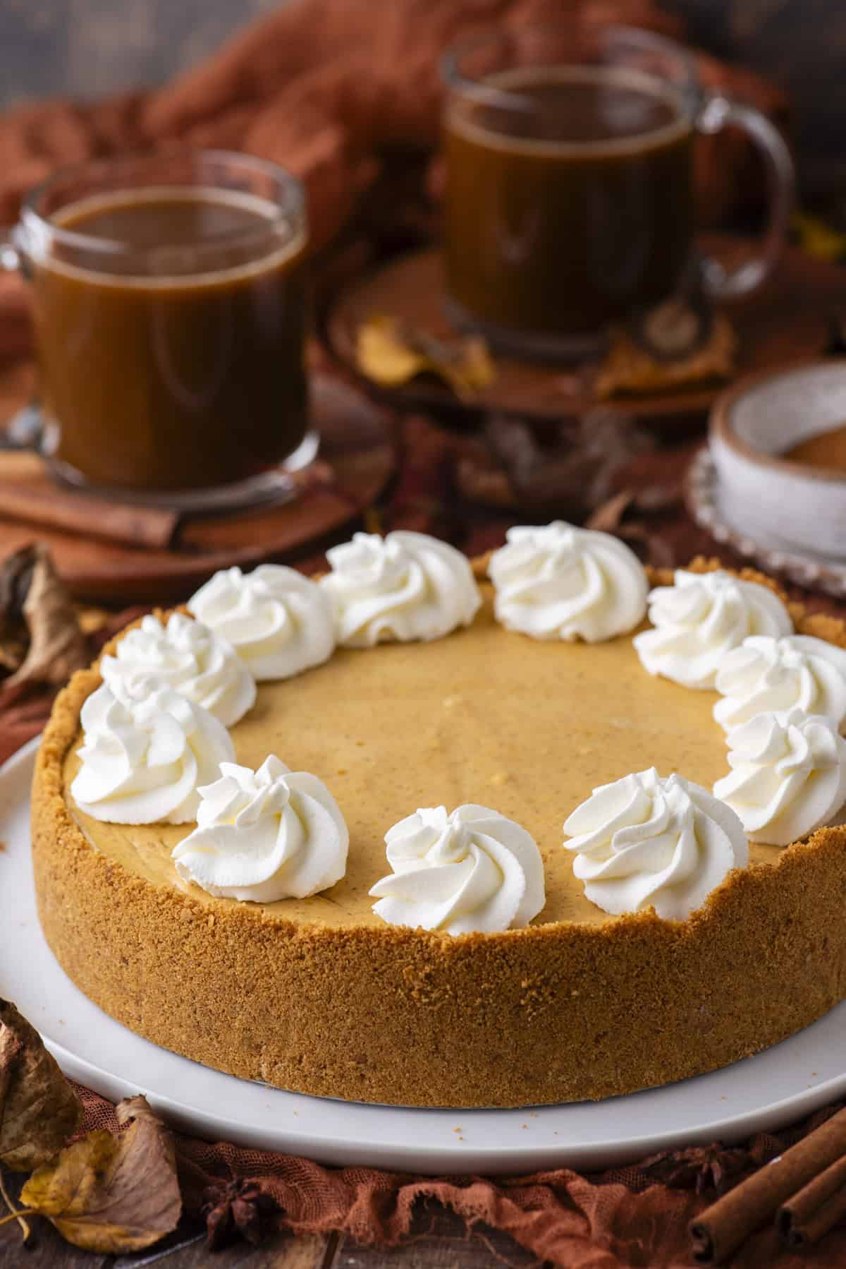 A pumpkin cheesecake with a graham cracker crust, topped with swirls of whipped cream, sits on a white plate. Two cups of coffee and autumn leaves are in the background.