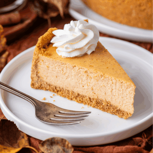 A slice of pumpkin cheesecake with a graham cracker crust, topped with a swirl of whipped cream, sits on a white plate with a fork beside it. Autumn leaves are scattered around the plate.