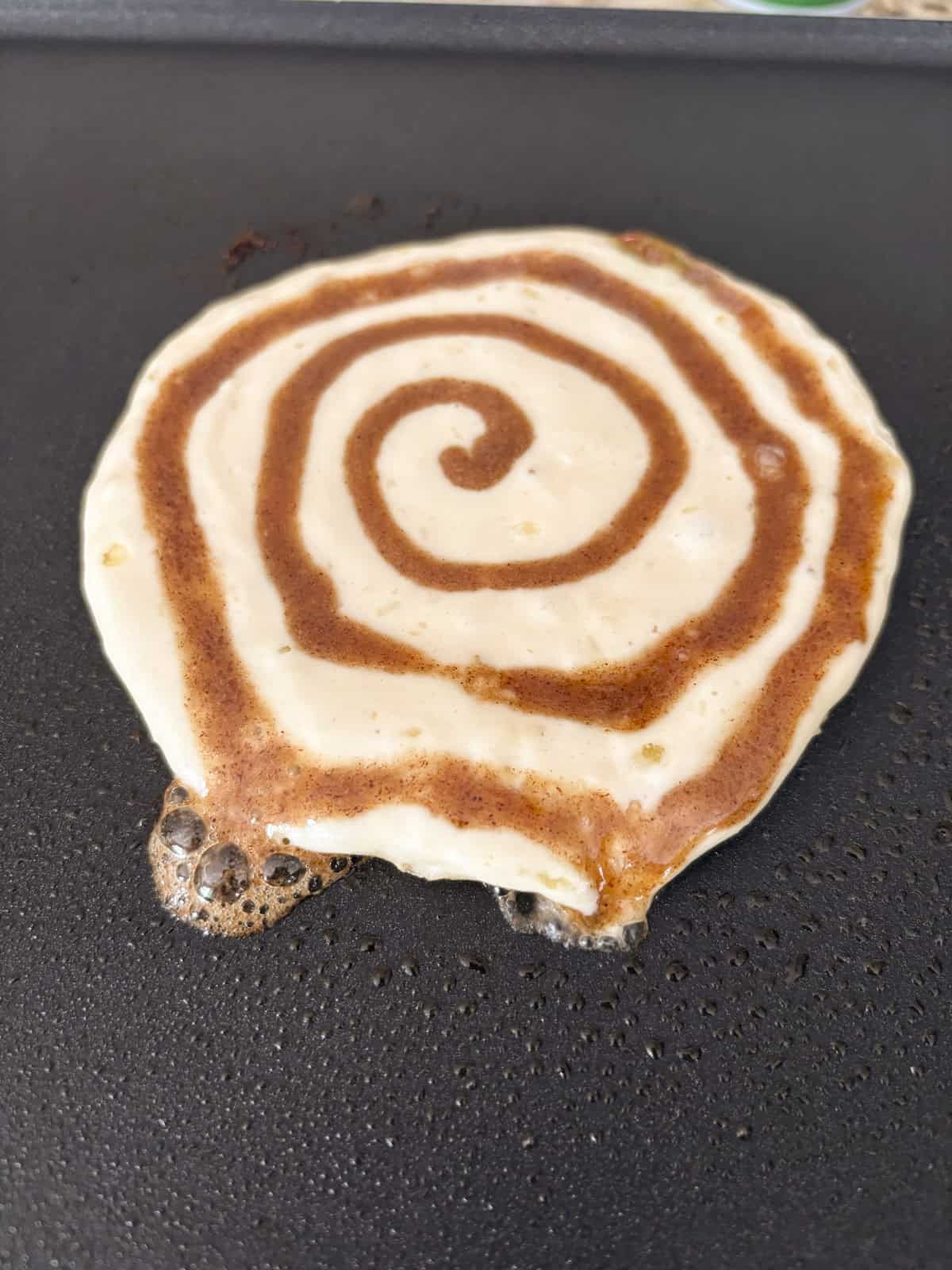 A pancake cooking on a griddle, with a visible cinnamon swirl pattern on top. Some bubbles are forming on the edge of the pancake as it cooks.